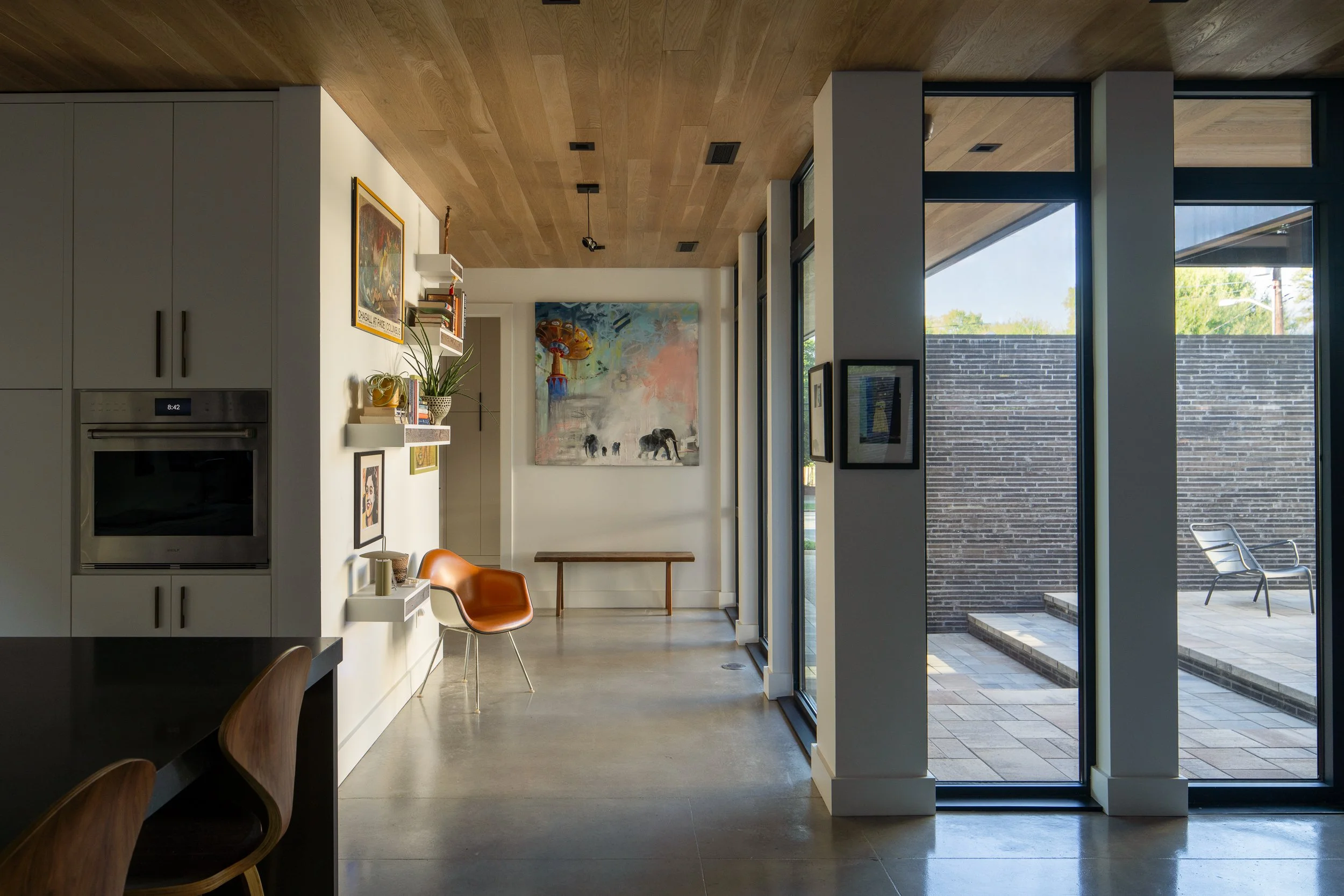 A wood-ceilinged interior passage with artwork and an orange chair opens toward a courtyard through full-height glazing.