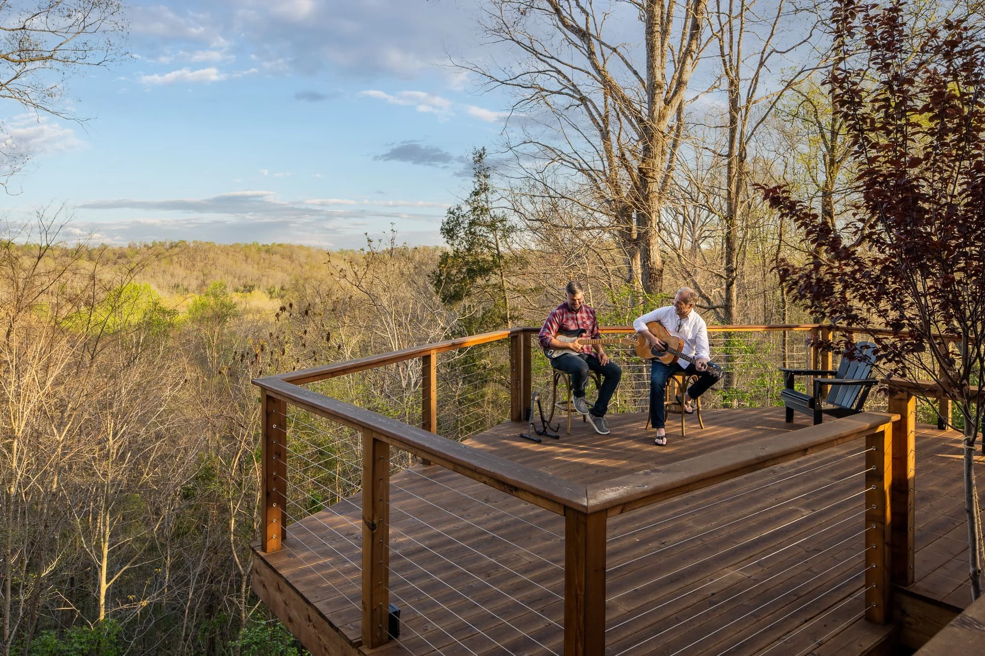 Two men sitting on a wooden deck, playing guitars, overlooking a wooded landscape with trees and a blue sky.