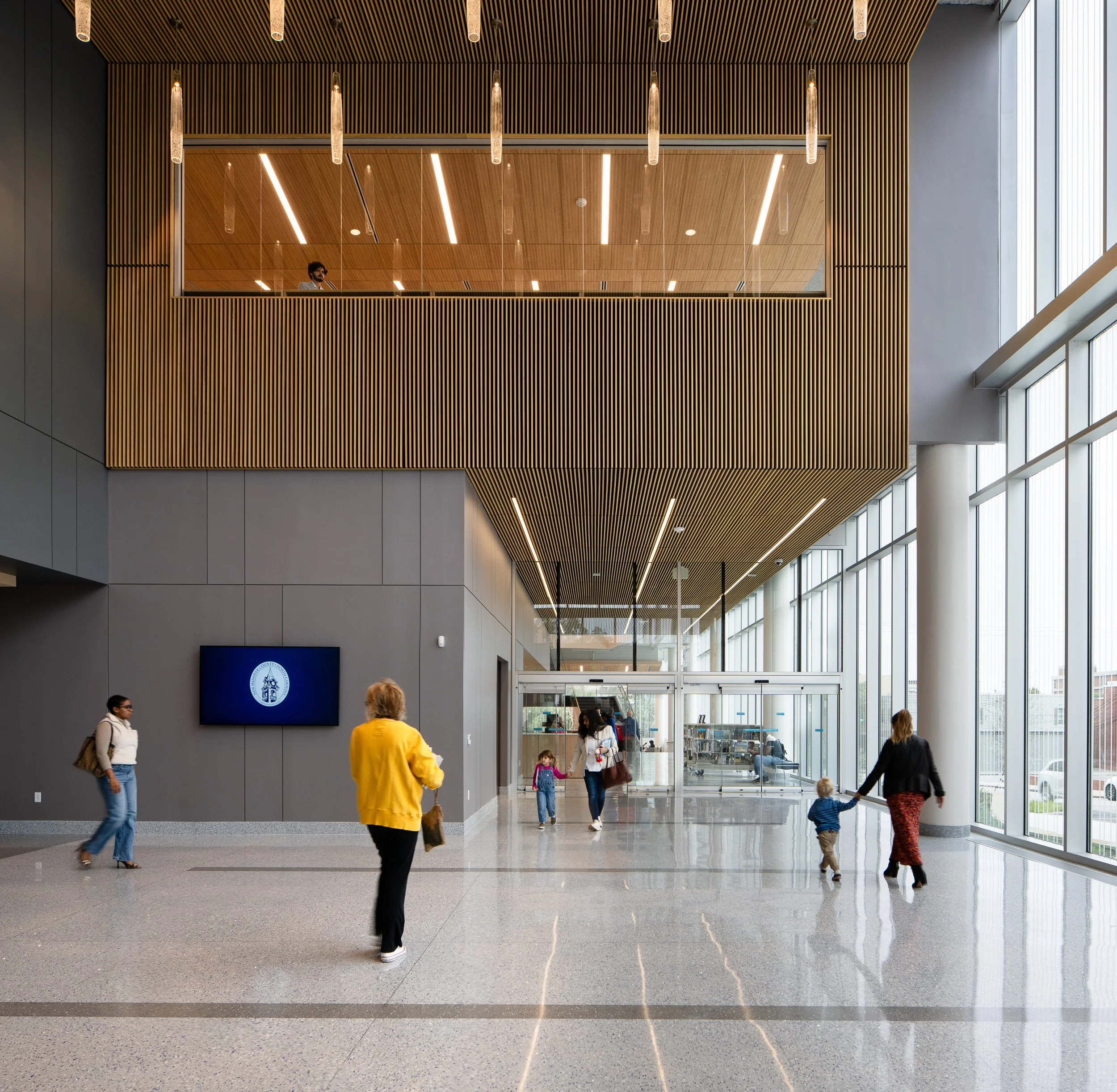 Main lobby with polished floors, tall windows, wood slat ceiling, pendant lights, and visitors.