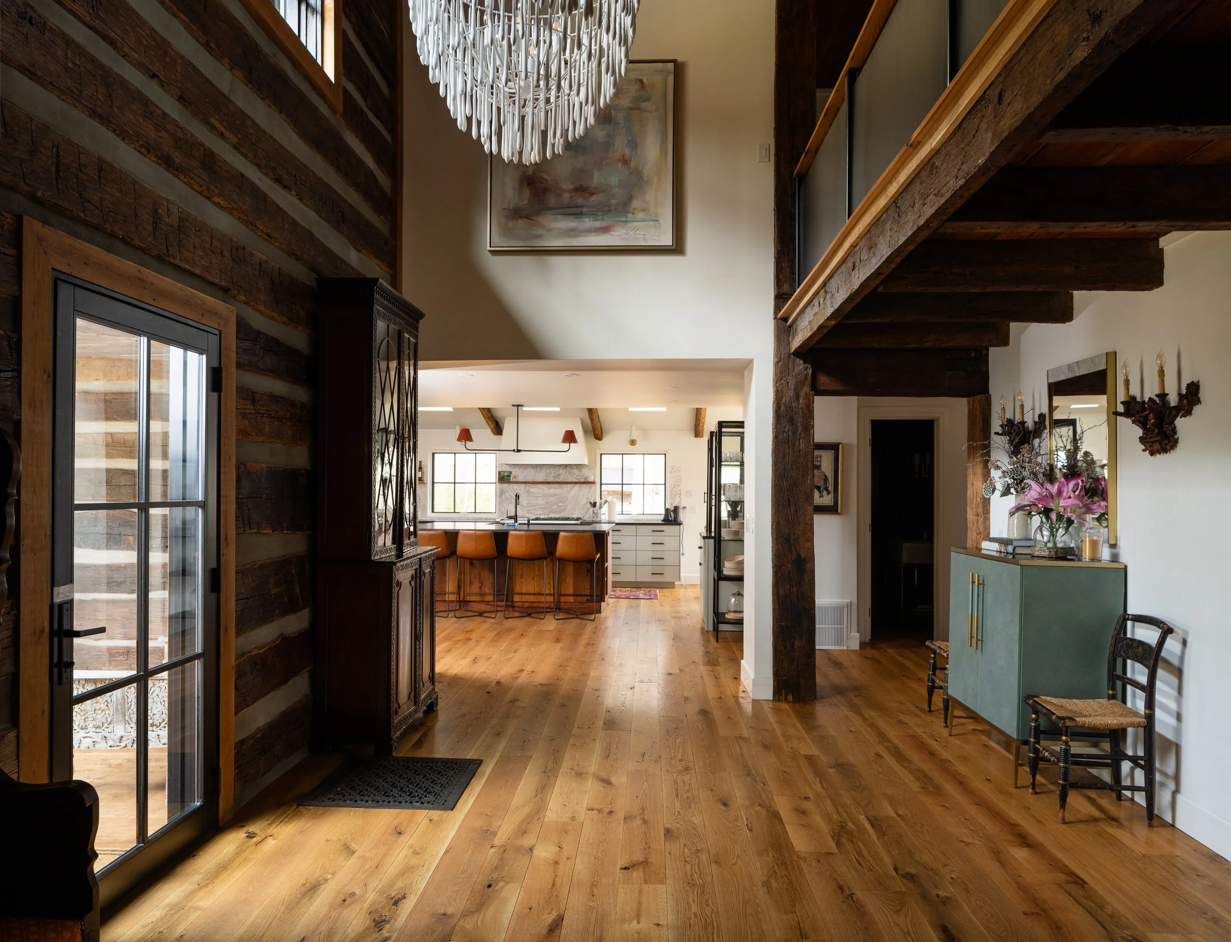 Entry hall with log walls, wide wood floors, exposed beams, chandelier, and a view into the kitchen.