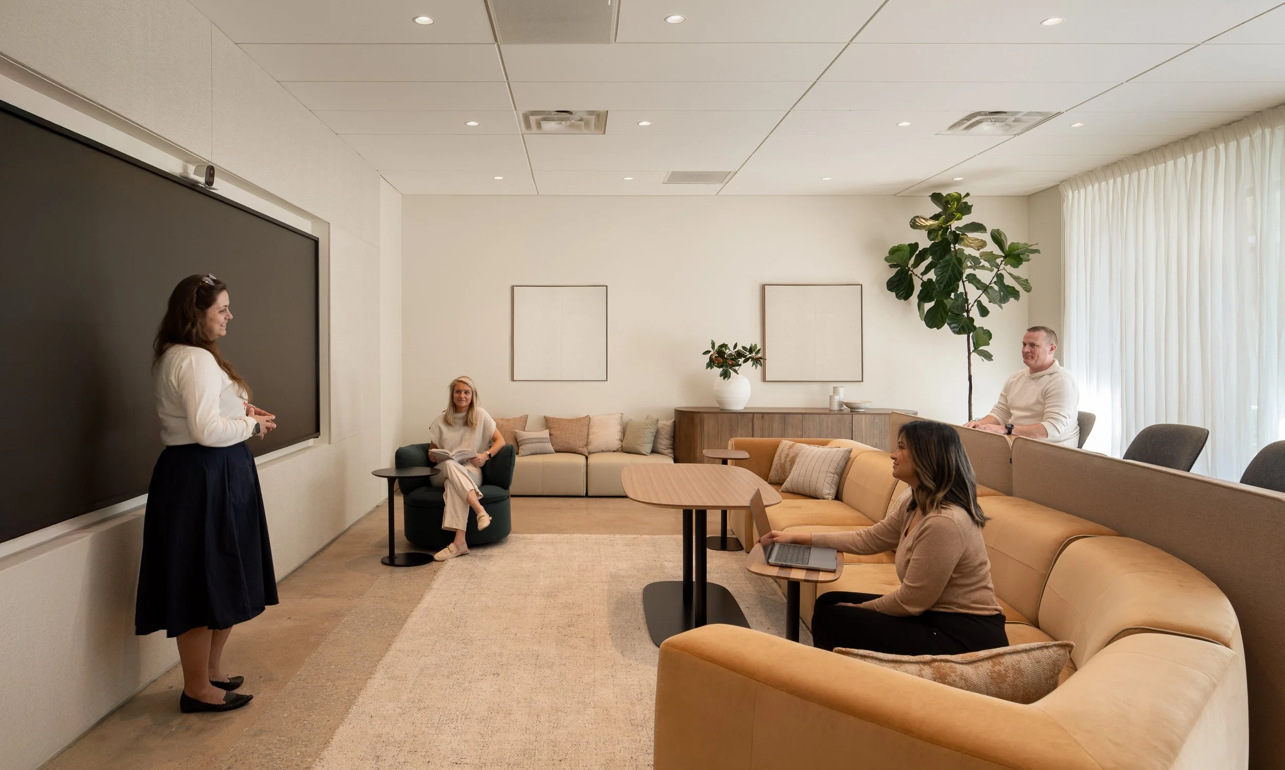 Team meeting room with a large mounted display, mixed seating including a tan modular sofa, green swivel chair, and dark sofa, plus a fiddle leaf fig and curtained window for diffused light.