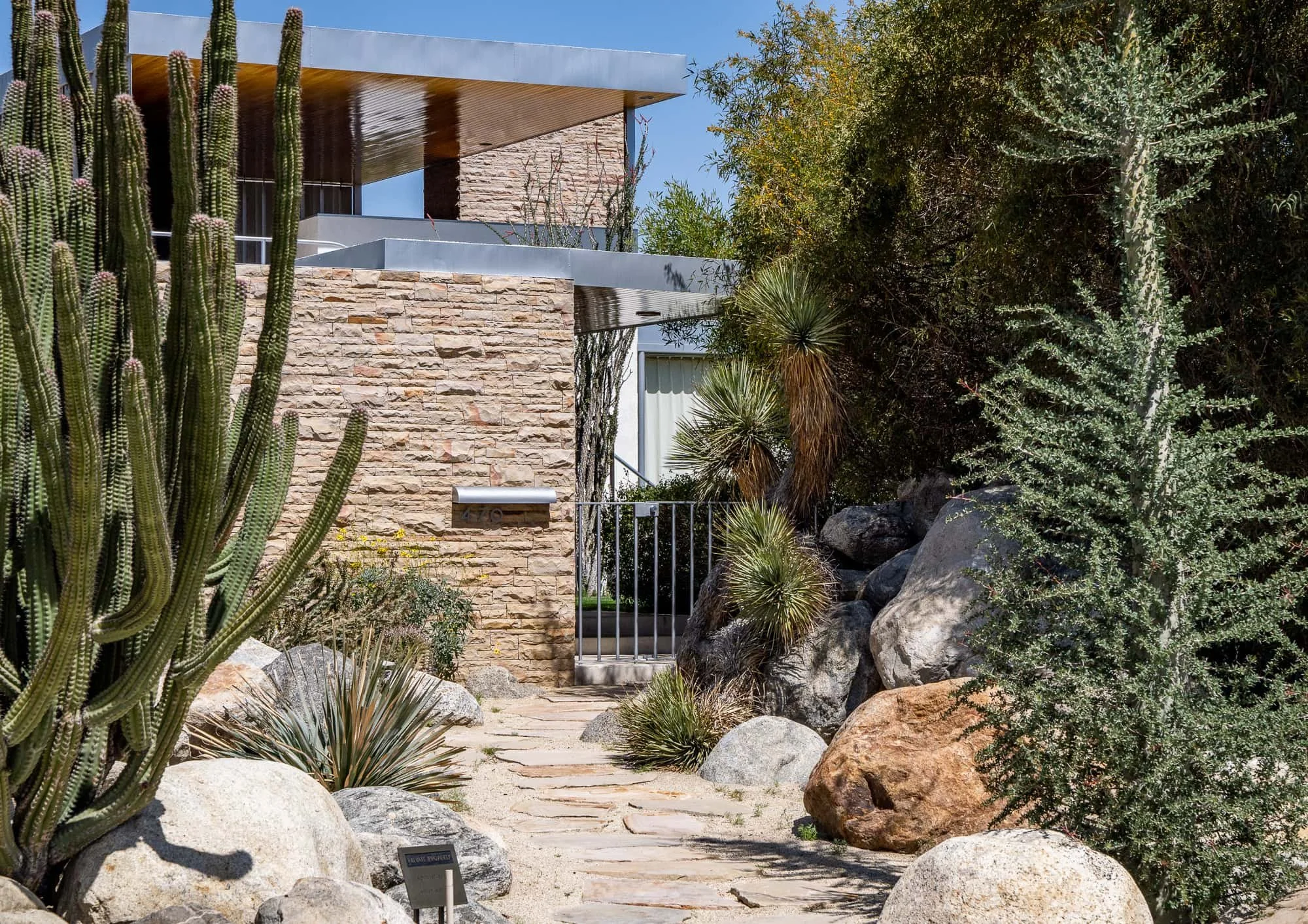 Residential walkway with stucco columns, palm trees, flowering plants, and layered desert landscaping.