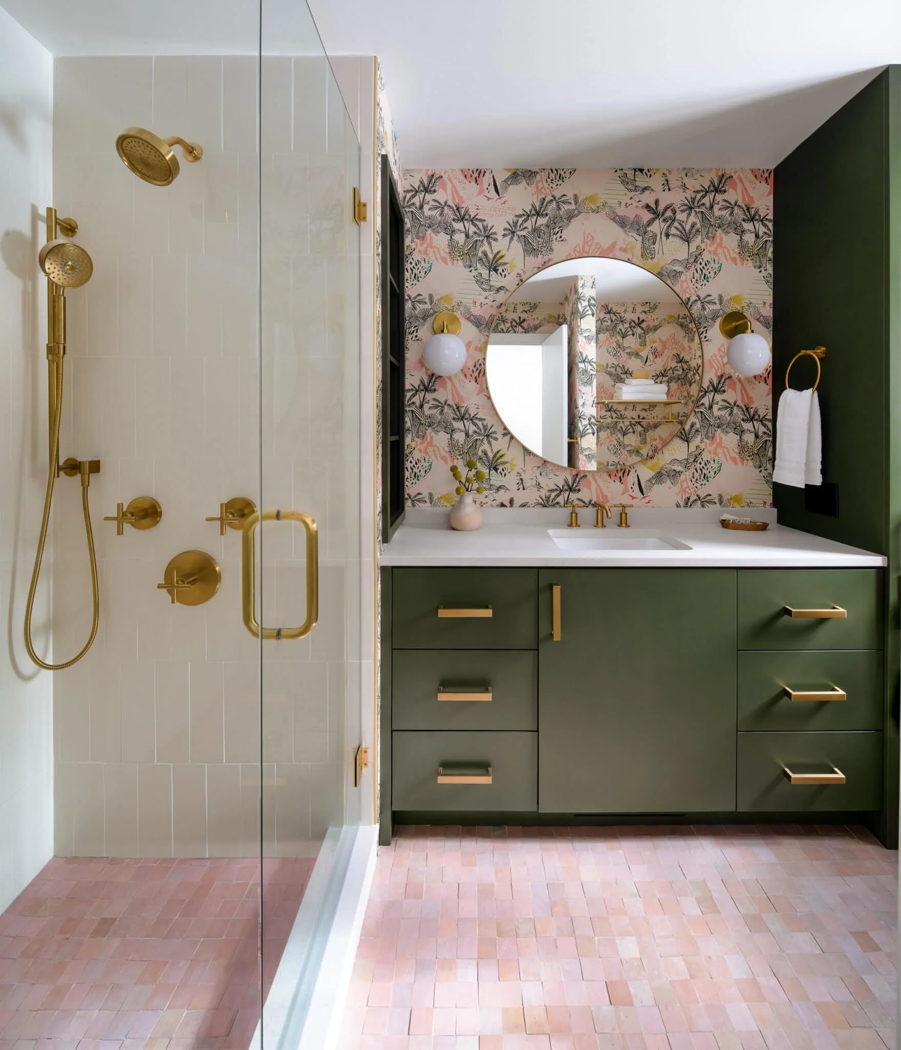Primary bathroom with dark green vanity, brass fixtures, floral wallpaper, round mirror, glass shower with white tile, and pink zellige floor tile