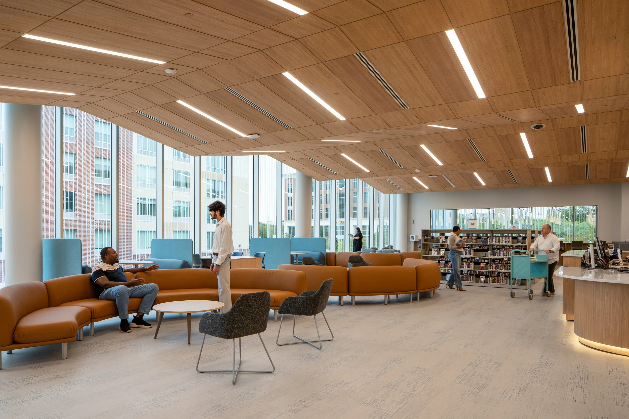 Modern library or lounge area with large windows, wooden ceiling, and seating. Several people are present, including a seated man, a standing man, a woman reading, and a man at a counter with a cart.