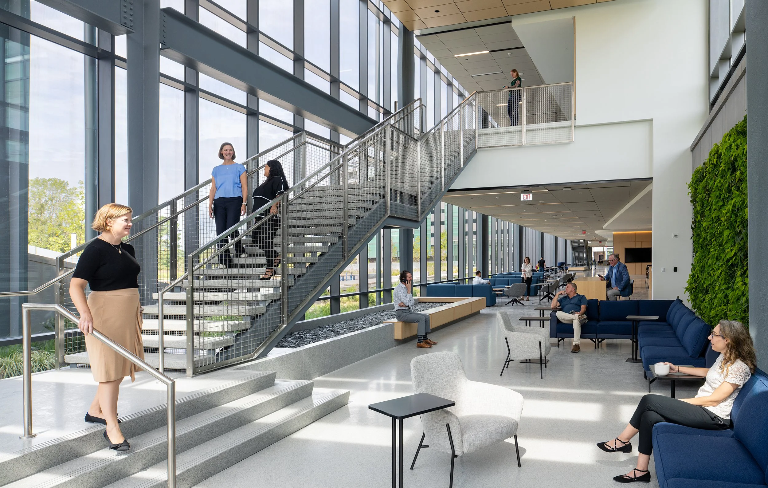 Open mesh-railed stair connecting the atrium floors, with staff moving between levels, a living wall on the right, and blue lounge seating along the curtain wall.