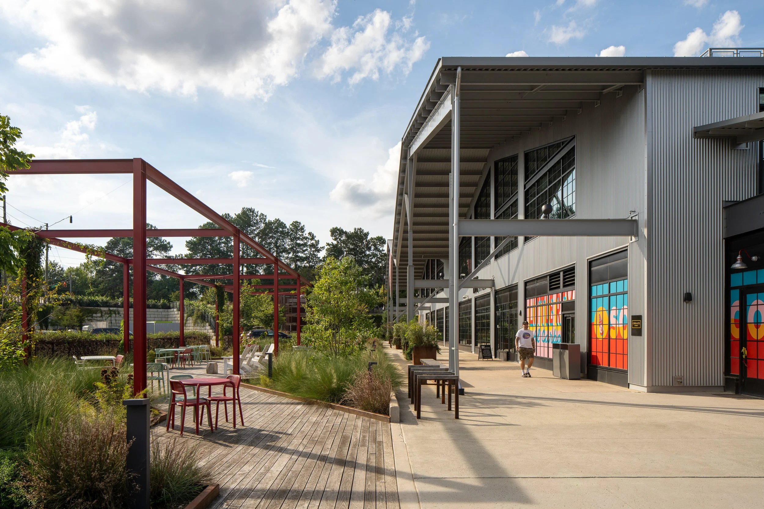 Outdoor courtyard with a wood boardwalk threading through native grasses, red steel pergola framing on the left, and the metal-clad retail building with colorful graphic storefronts on the right.