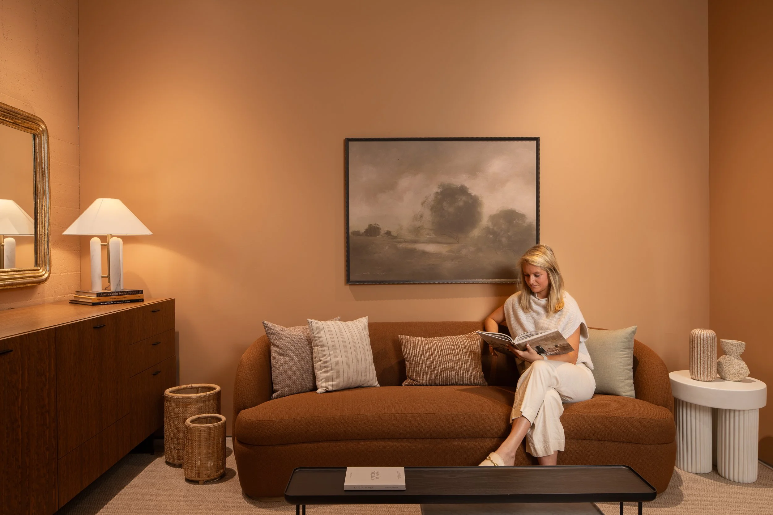 Small lounge with warm terracotta walls, a curved cognac sofa, a dark wood console with table lamp and gilt mirror, and a moody landscape painting hung above the sofa.