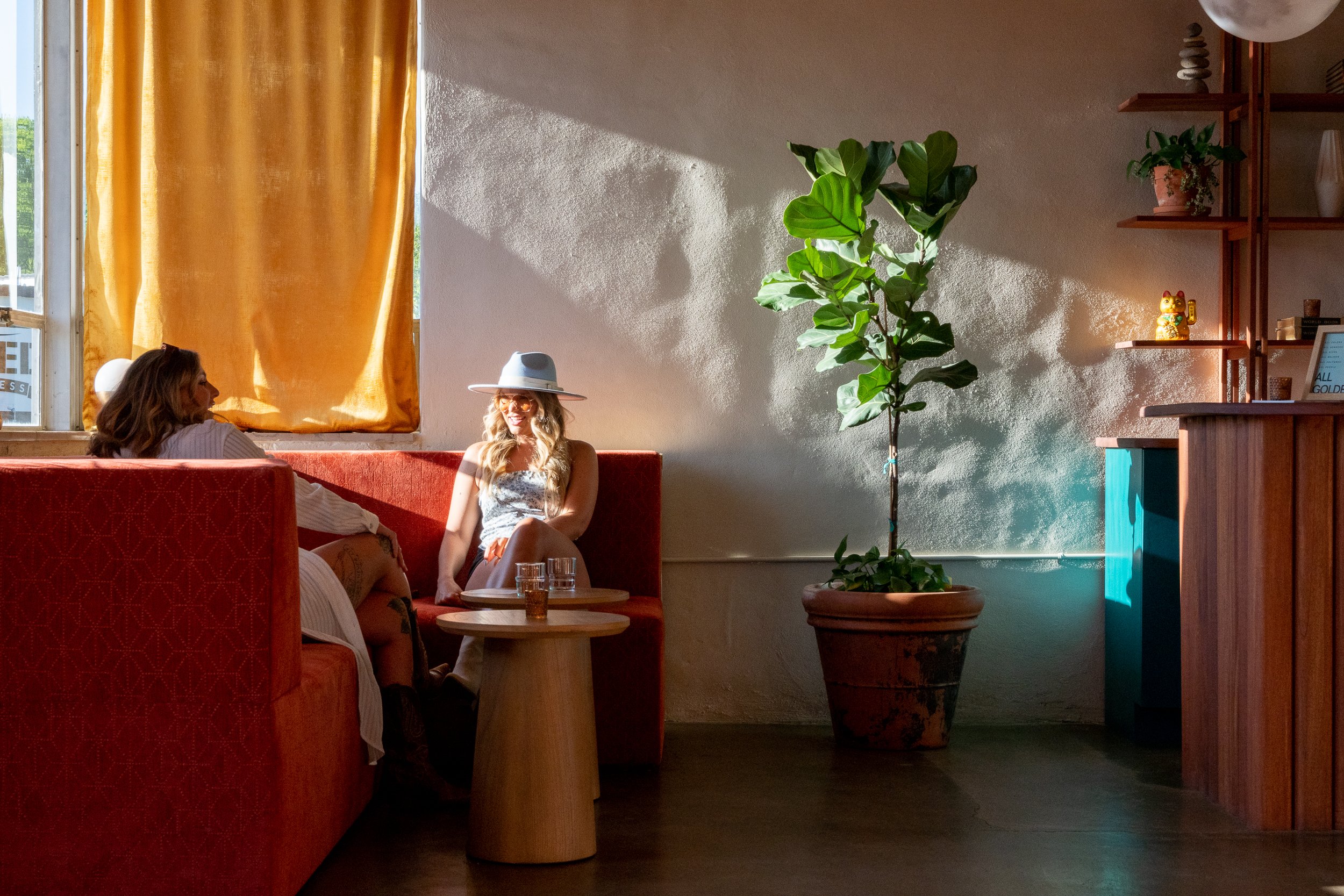 Two women conversing in a warm-lit lounge with red booth seating and a potted plant.