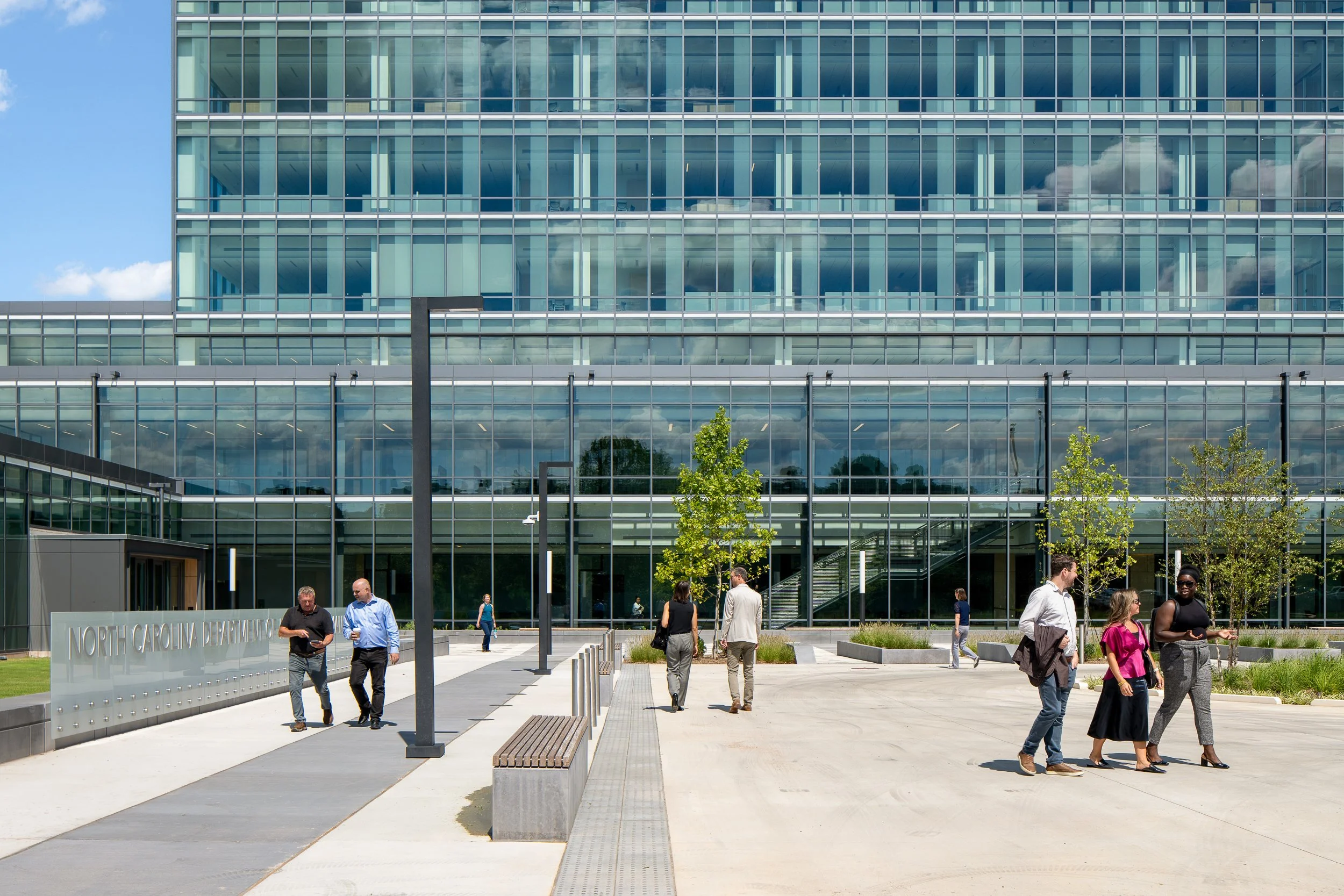 Plaza view toward the main entrance with visitors walking past benches and young trees, framed against the reflective glass curtain wall.