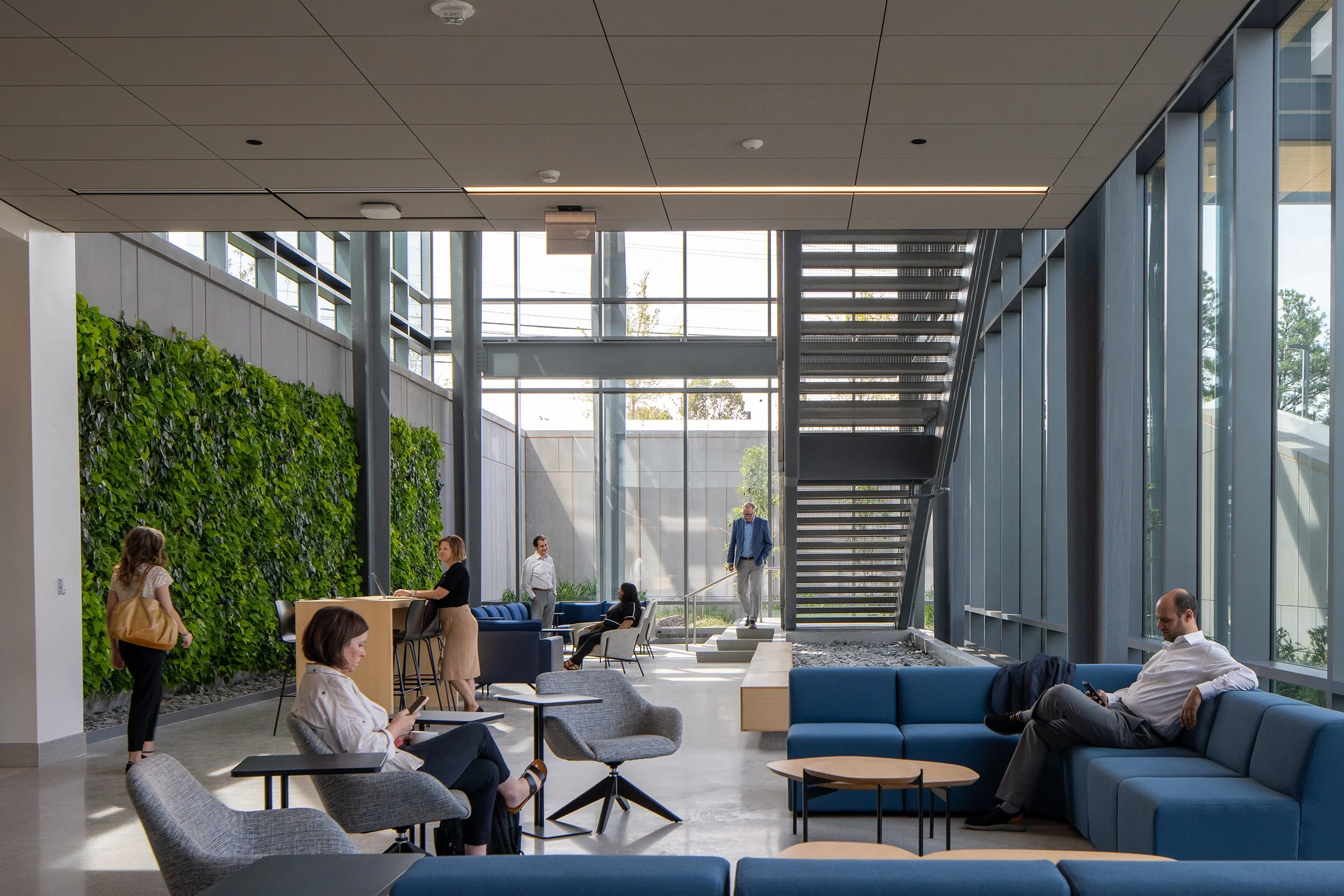 Interior lounge with a full-height living wall on the left, modern blue and gray seating, an exposed steel stair on the right, and daylight pouring through the curtain wall.