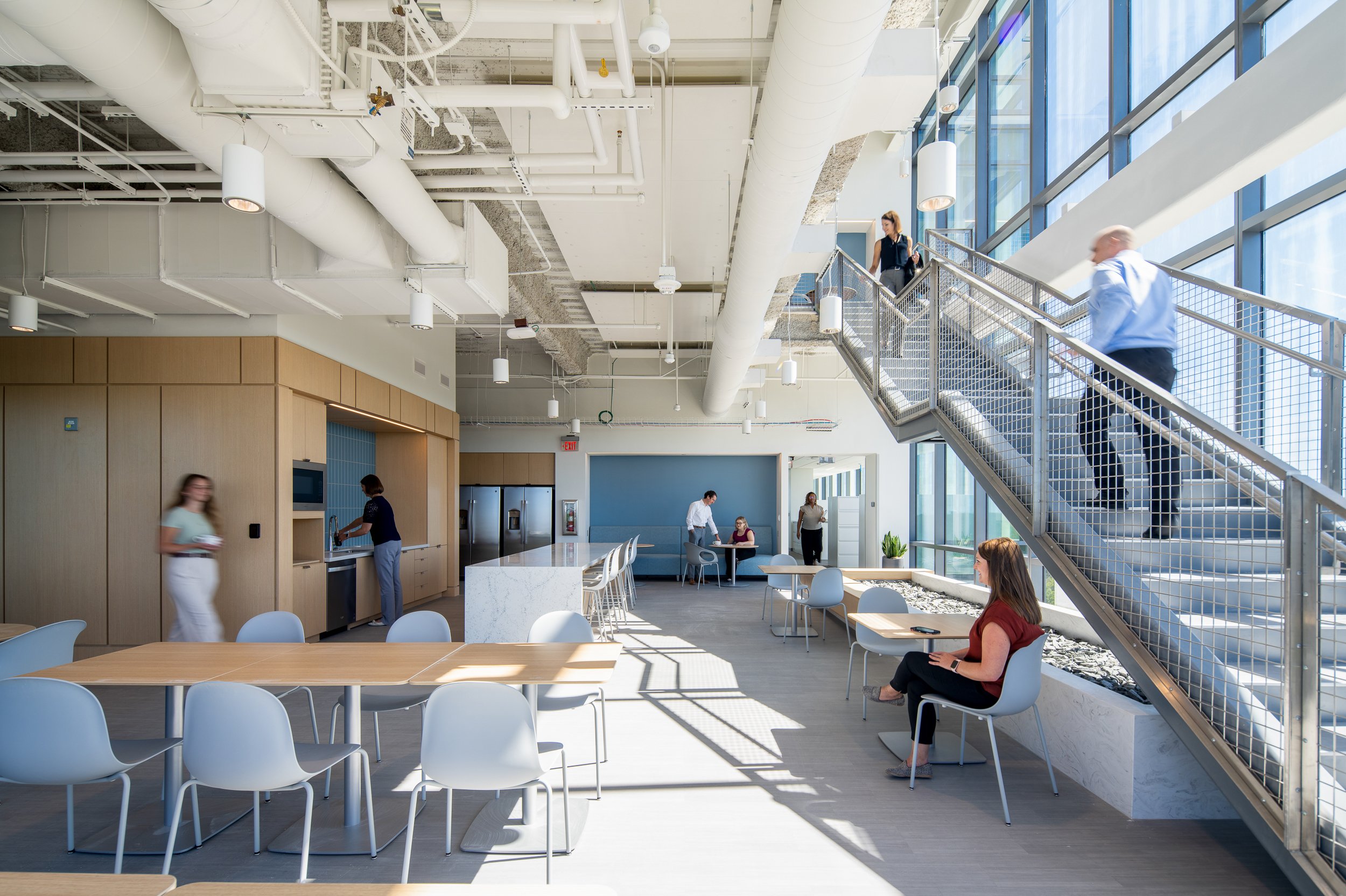 Break and dining area beneath an open mesh-railed stair, with exposed white ductwork above, a wood millwork kitchenette, and staff moving between tables and the staircase.
