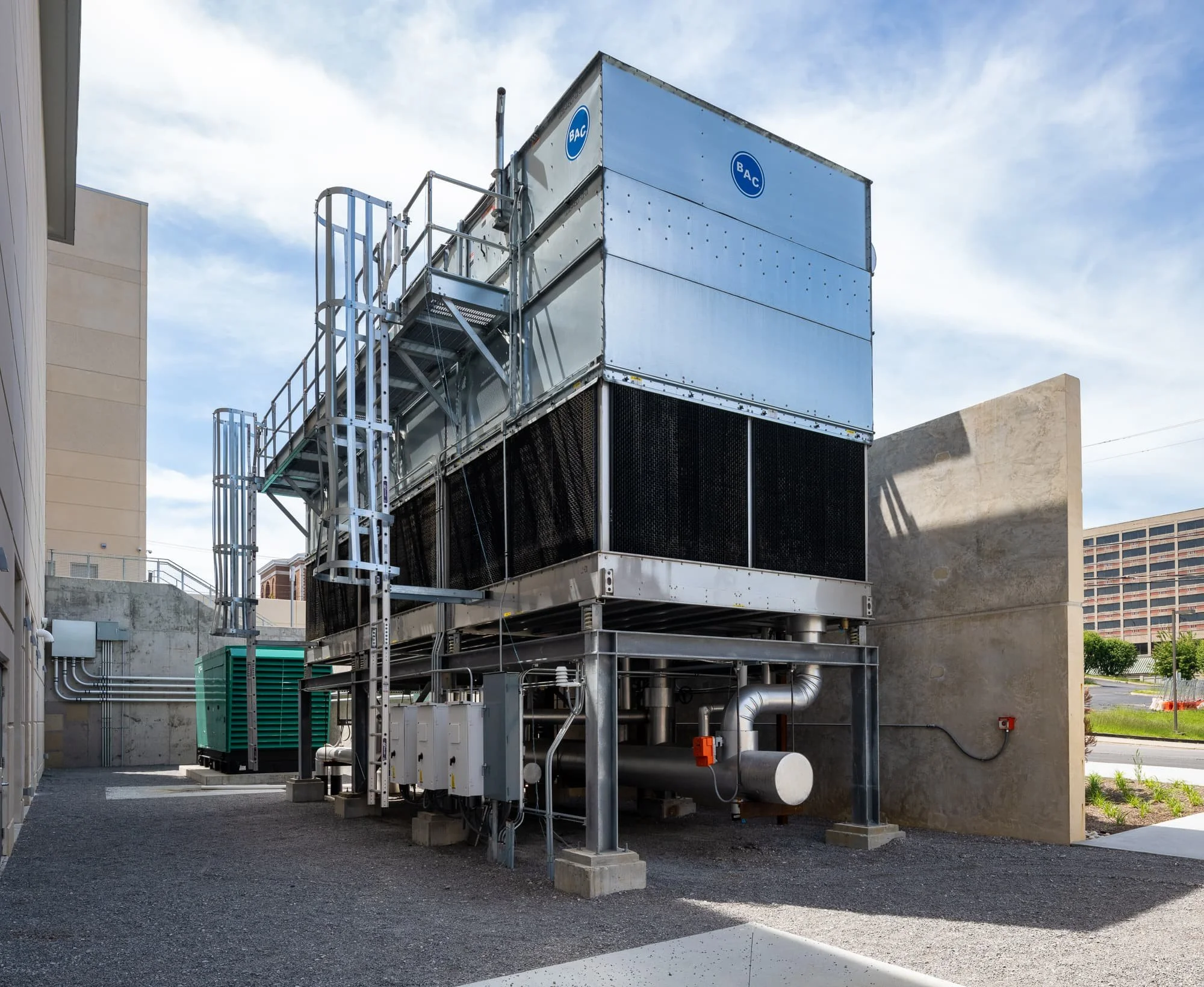 Outdoor mechanical equipment area with a large cooling tower, metal access ladders, concrete service walls, and utility piping.