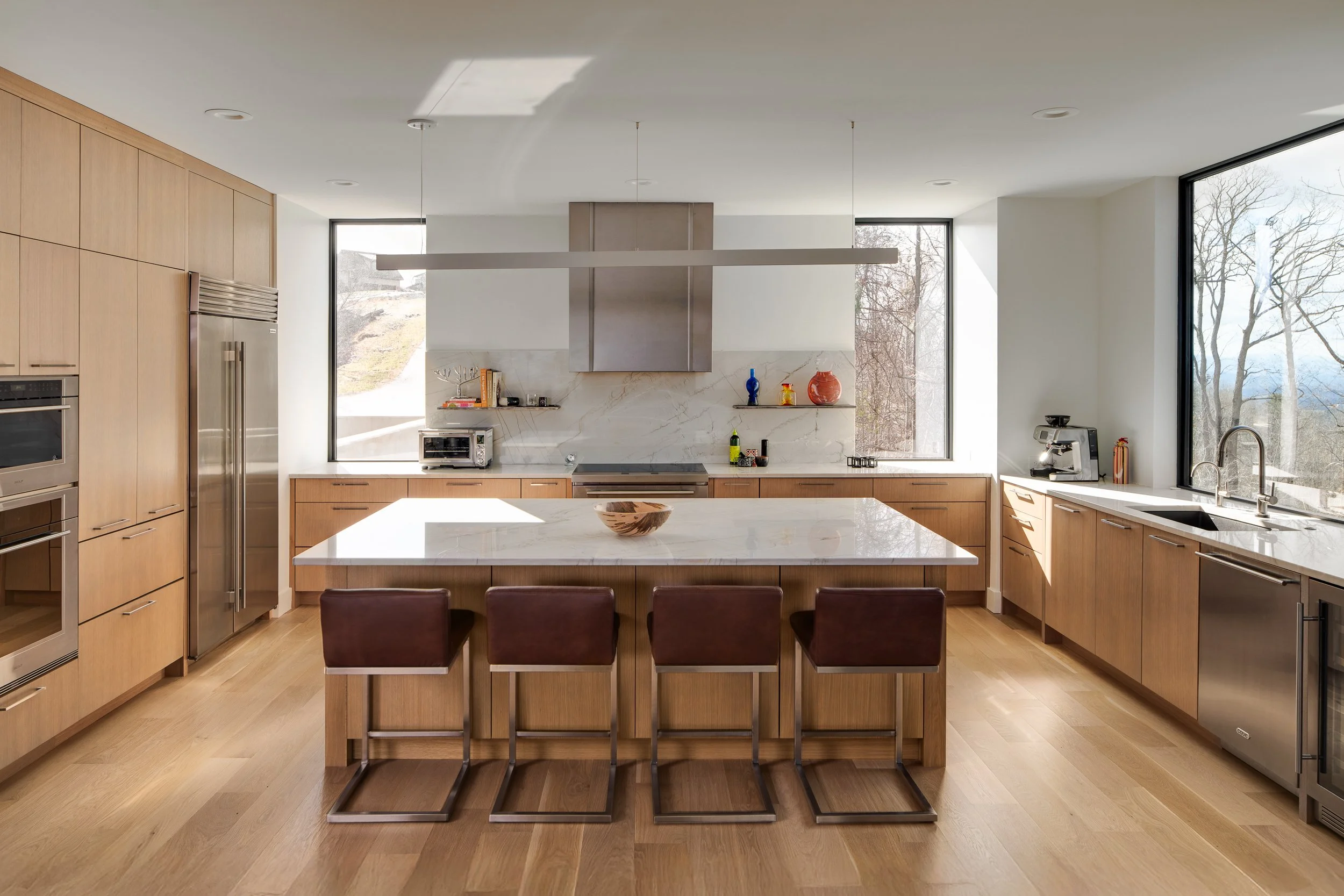 Bright modern kitchen with pale wood cabinetry, marble island, stainless appliances, and large windows.