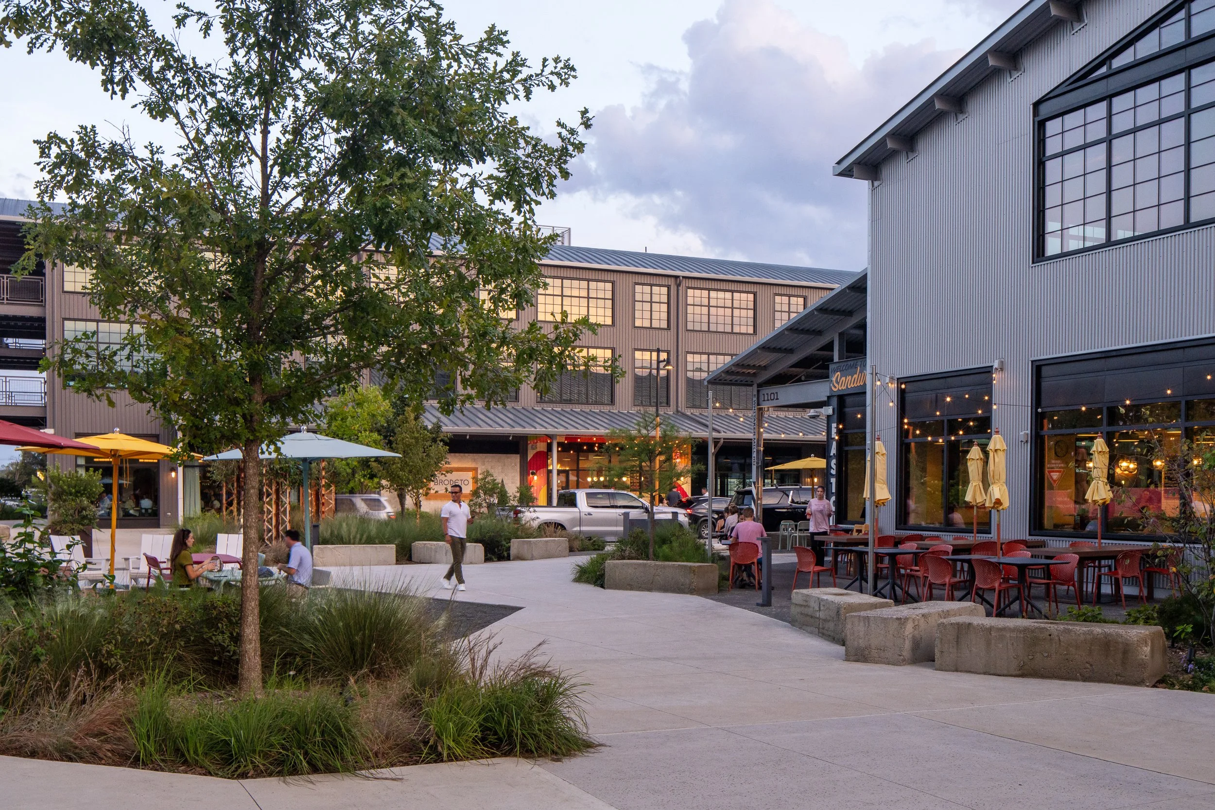 Plaza at twilight with a young oak tree in the foreground, the Brodeto and Eastcut storefronts glowing from within, outdoor diners under string lights, and the metal-clad buildings against the evening sky.
