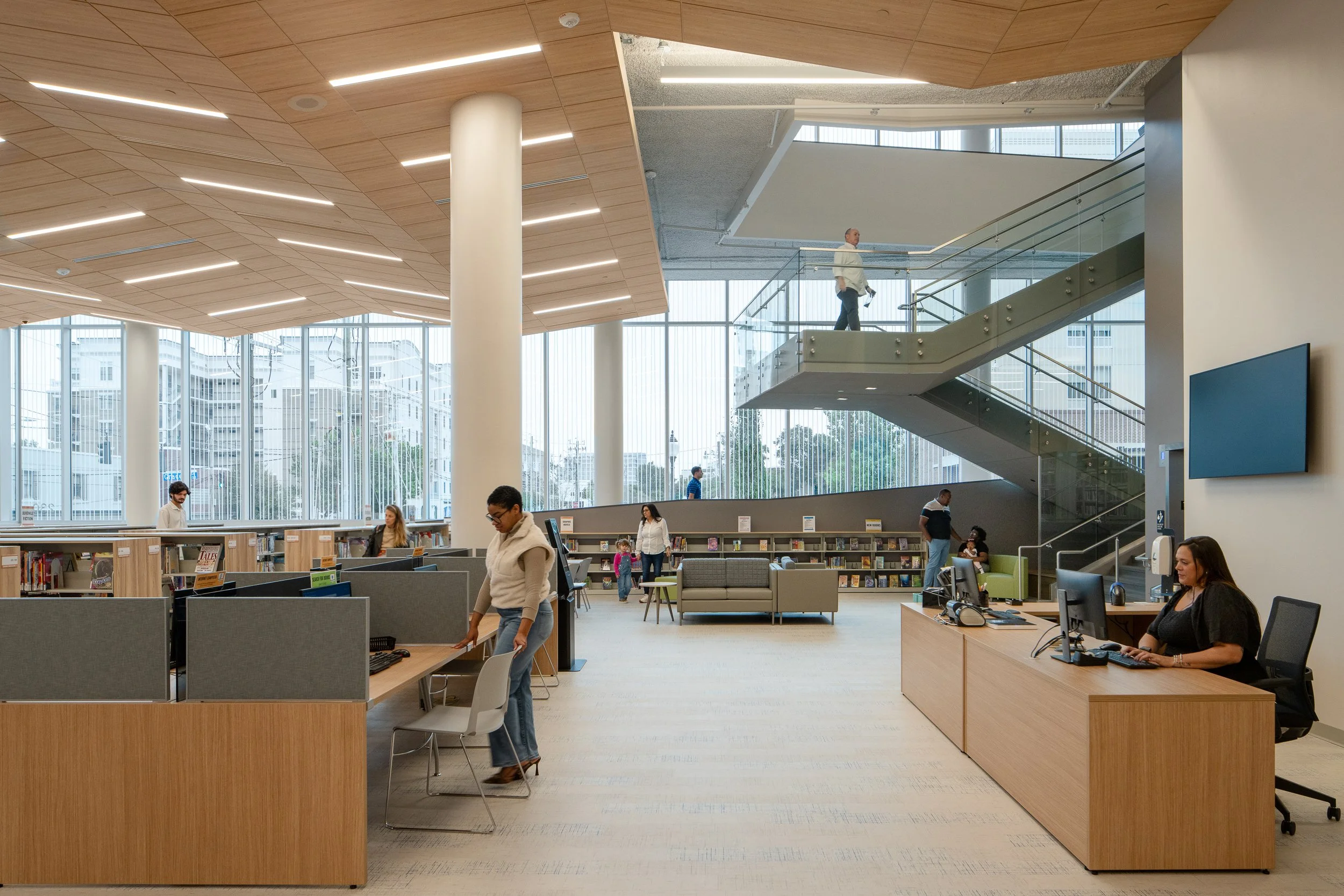 Interior service and reading area with wood ceilings, tall windows, public desks, and glass stair.