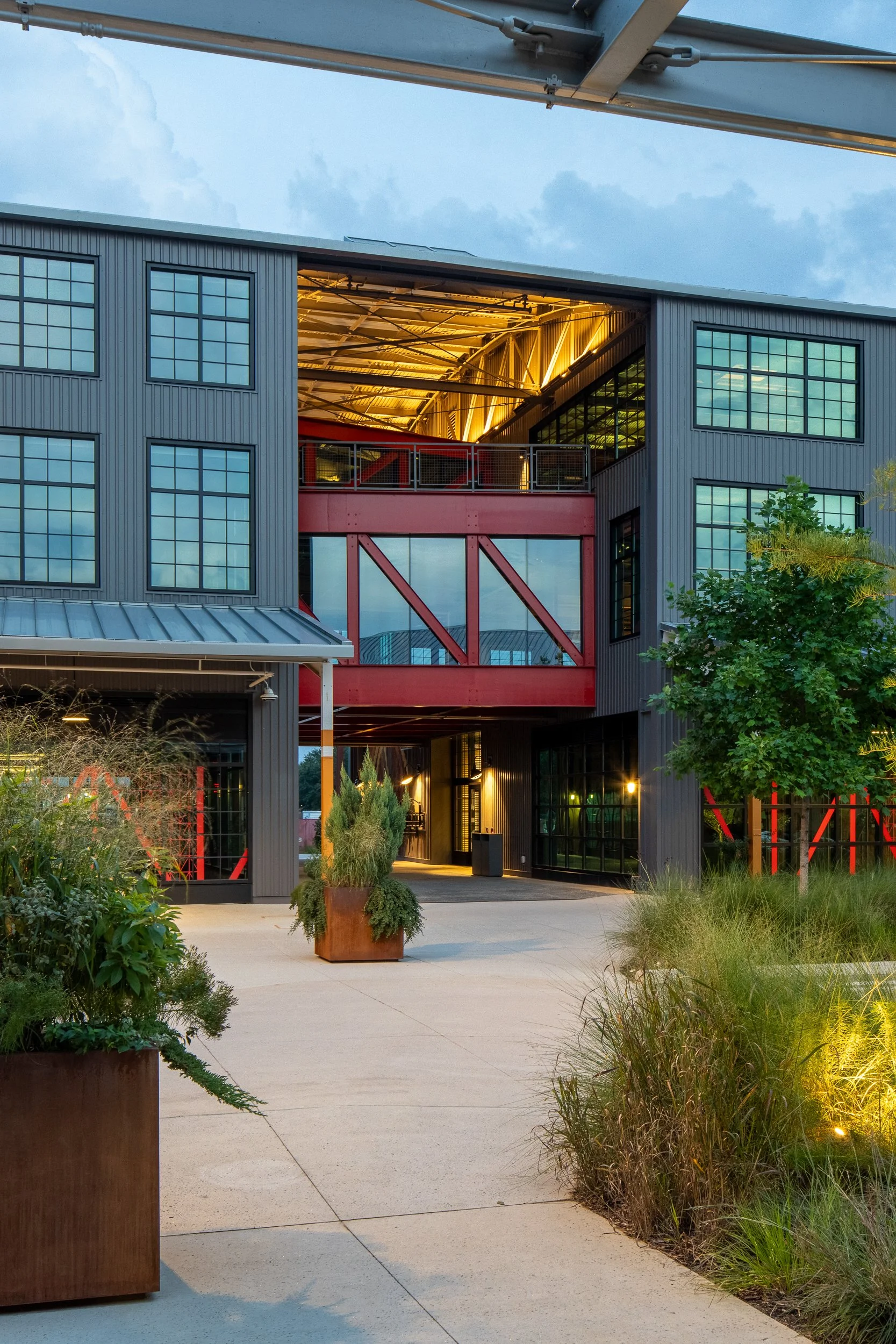 Twilight view of the central entrance with the red diagonal-braced steel bridge glowing against warm interior light, native grass plantings in the foreground, and the corrugated metal facade against a deep blue sky.
