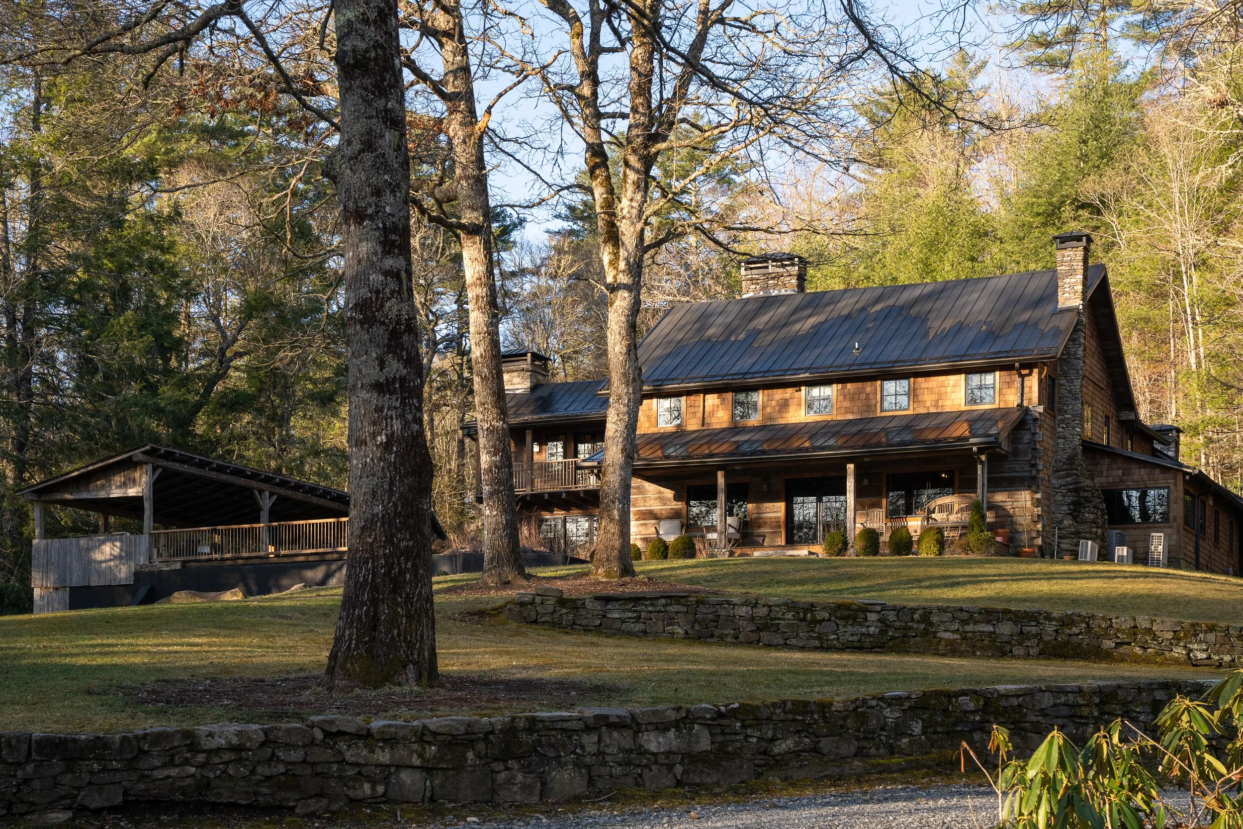 A large house surrounded by trees with leafless branches and green foliage, set in a landscaped yard with grass, a stone wall, and a gravel path, beneath a clear blue sky.