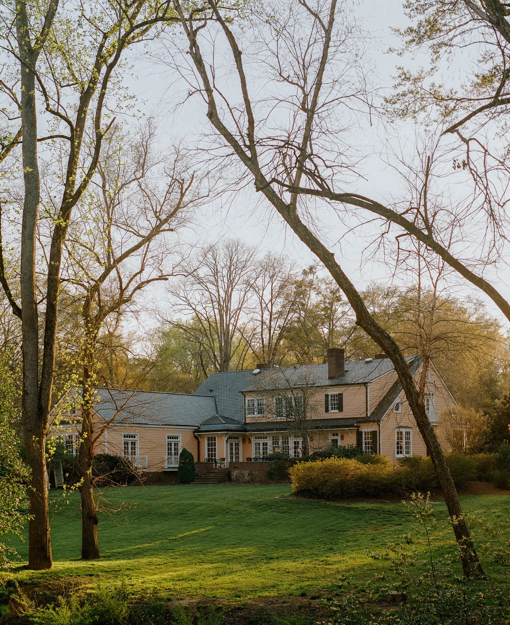 A large pink house with a gray roof, surrounded by trees and a well-maintained lawn, during daytime with sunlight filtering through the branches.