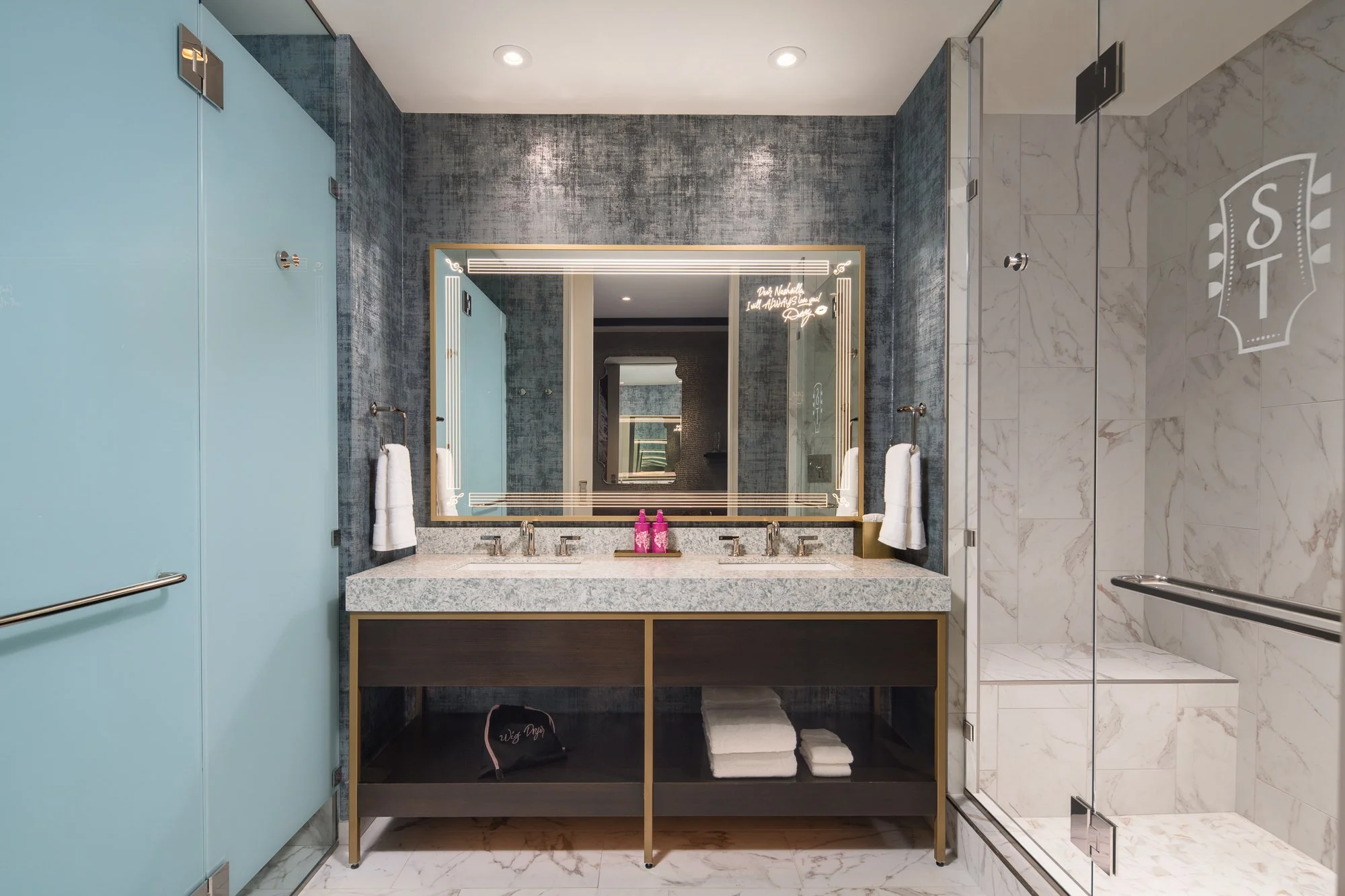 Guest bathroom at SongTeller Hotel featuring a double vanity, illuminated mirror, frosted glass enclosure, and marble shower.