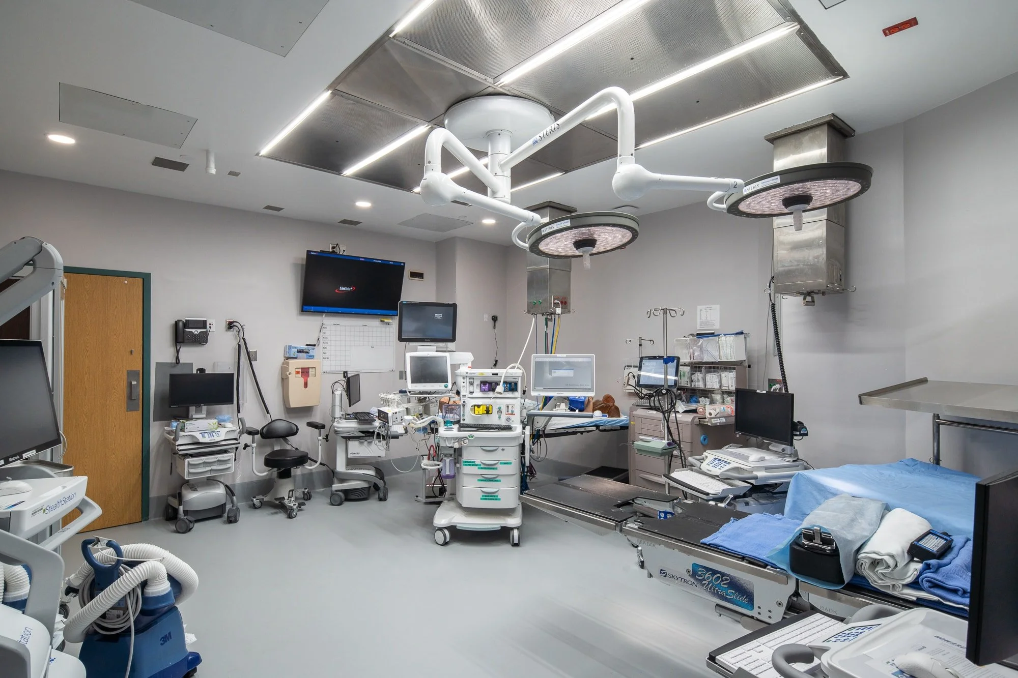 Modern hospital operating room with surgical lights, medical monitors, anesthesia equipment, stainless steel tables, and clinical workstations.