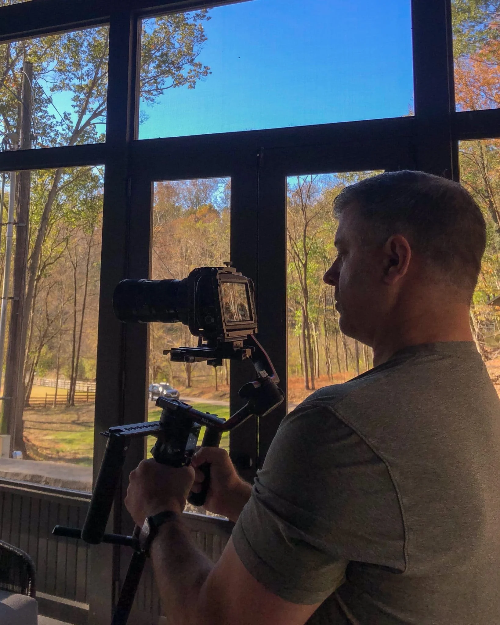 Man preparing a camera on a tripod indoors in front of a large window with trees outside, during daytime.