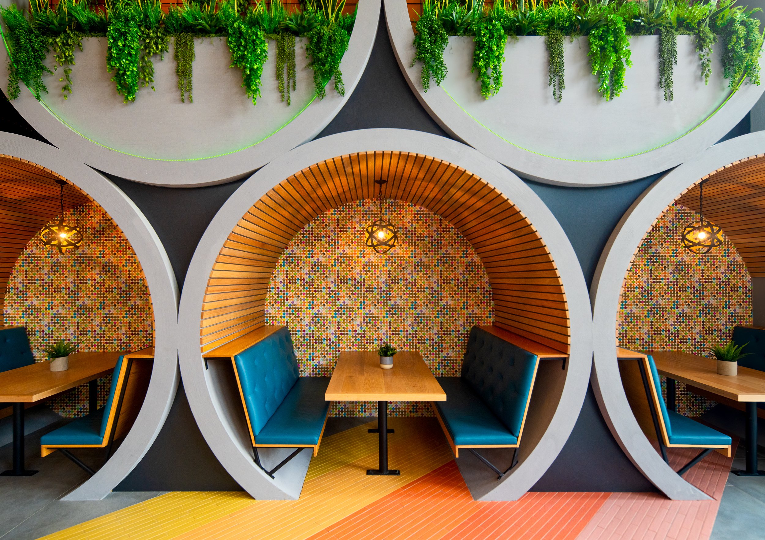 Interior of a modern restaurant with blue cushioned booths, wooden tables, and decorative circular walls with colorful mosaic patterns. Green plants and round hanging light fixtures are above the booths, and the ceiling features alternating curved structures with greenery.