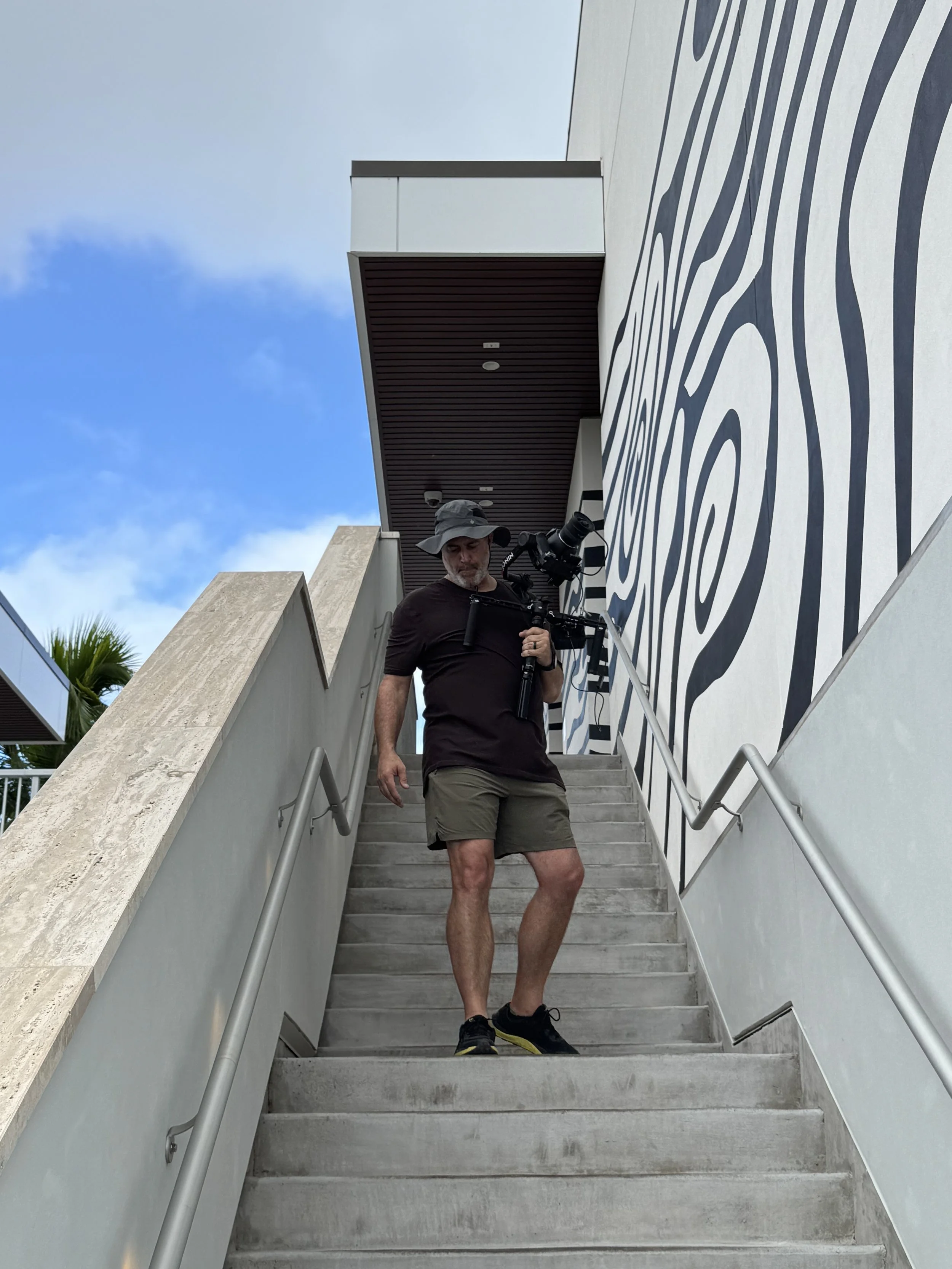 A man with a hat, beard, black shirt, and shorts standing on outdoor concrete stairs, holding a camera with a tripod, against a backdrop of a building with a black and white zebra stripe mural and a blue sky with clouds.