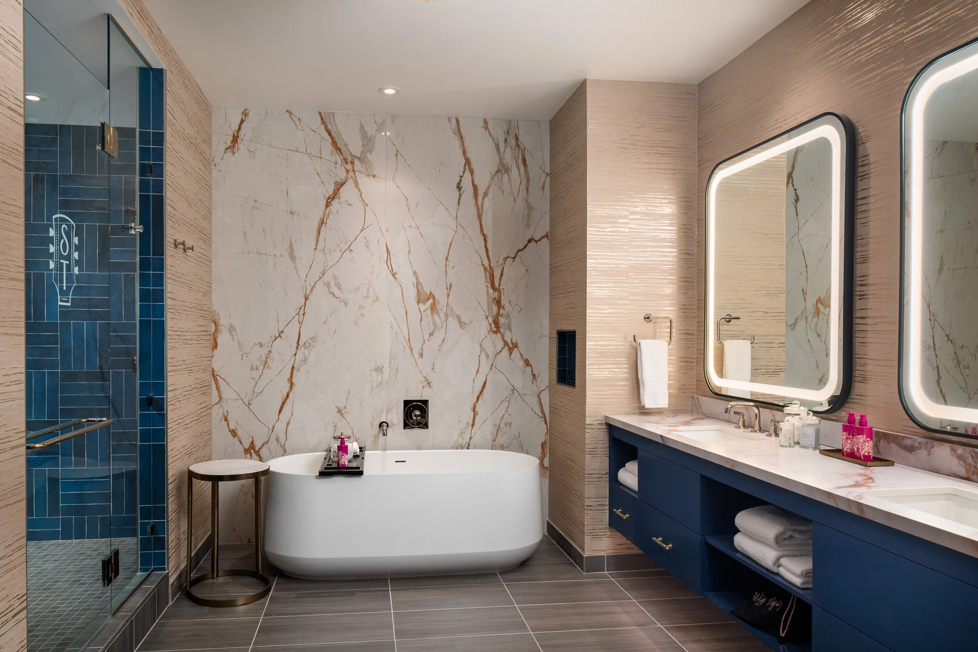 Luxury hotel bathroom with a freestanding soaking tub, dramatic marble wall, blue vanity, illuminated mirrors, and tiled shower.