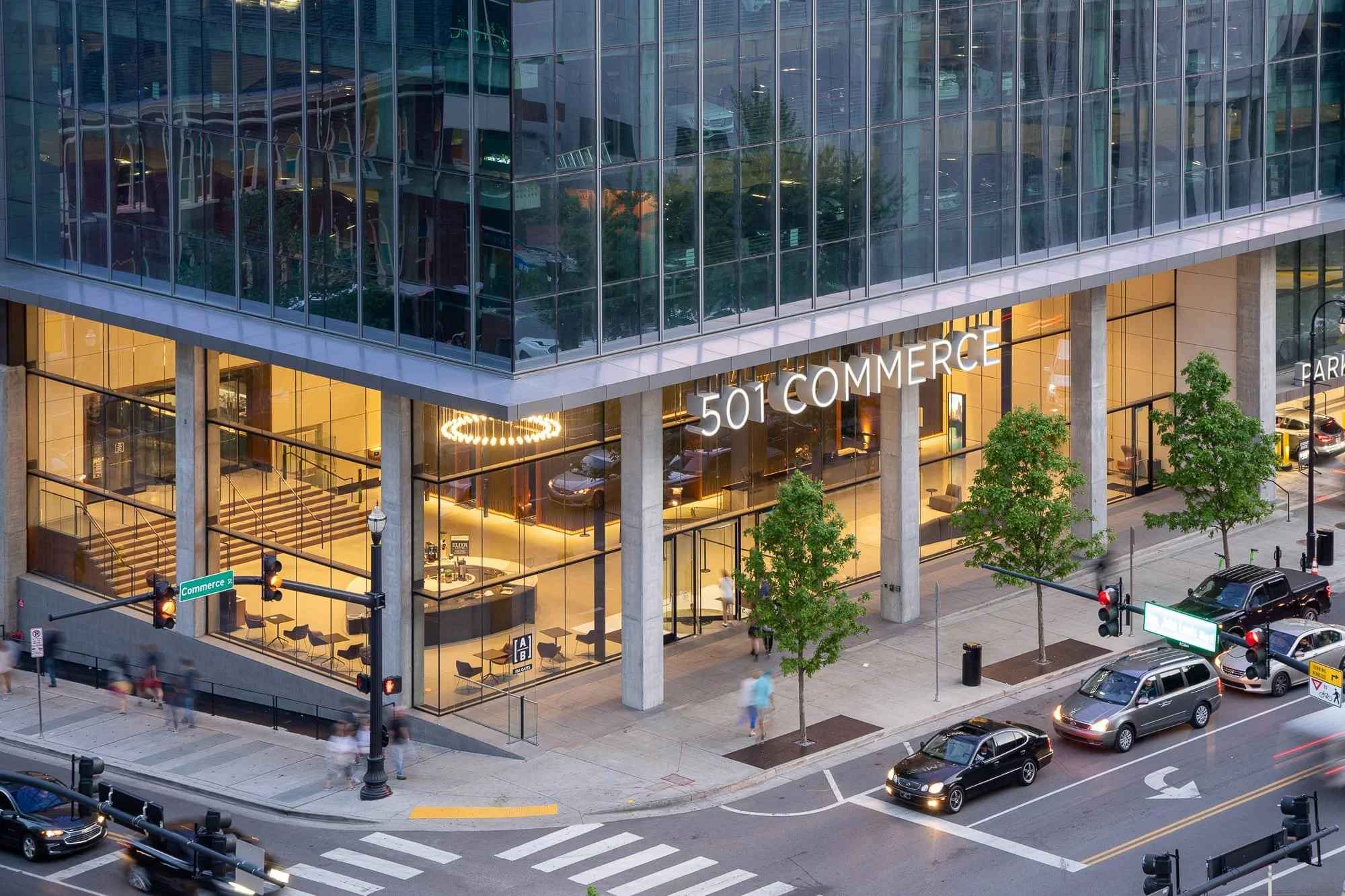 Street-level view of 501 Commerce with glass tower facade, lit lobby, crosswalks, and downtown traffic.