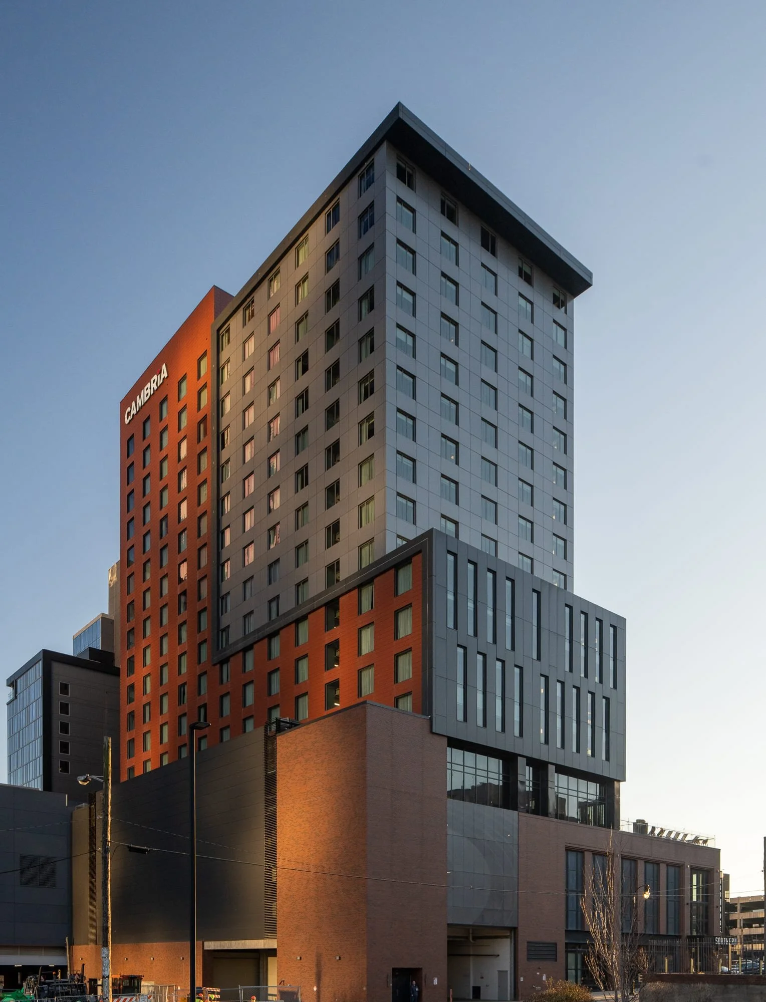 Side by side of the Cambria Hotel rear facade in evening showing dappled reflected light from surrounding buildings creating organic shadow patterns across the gray cladding