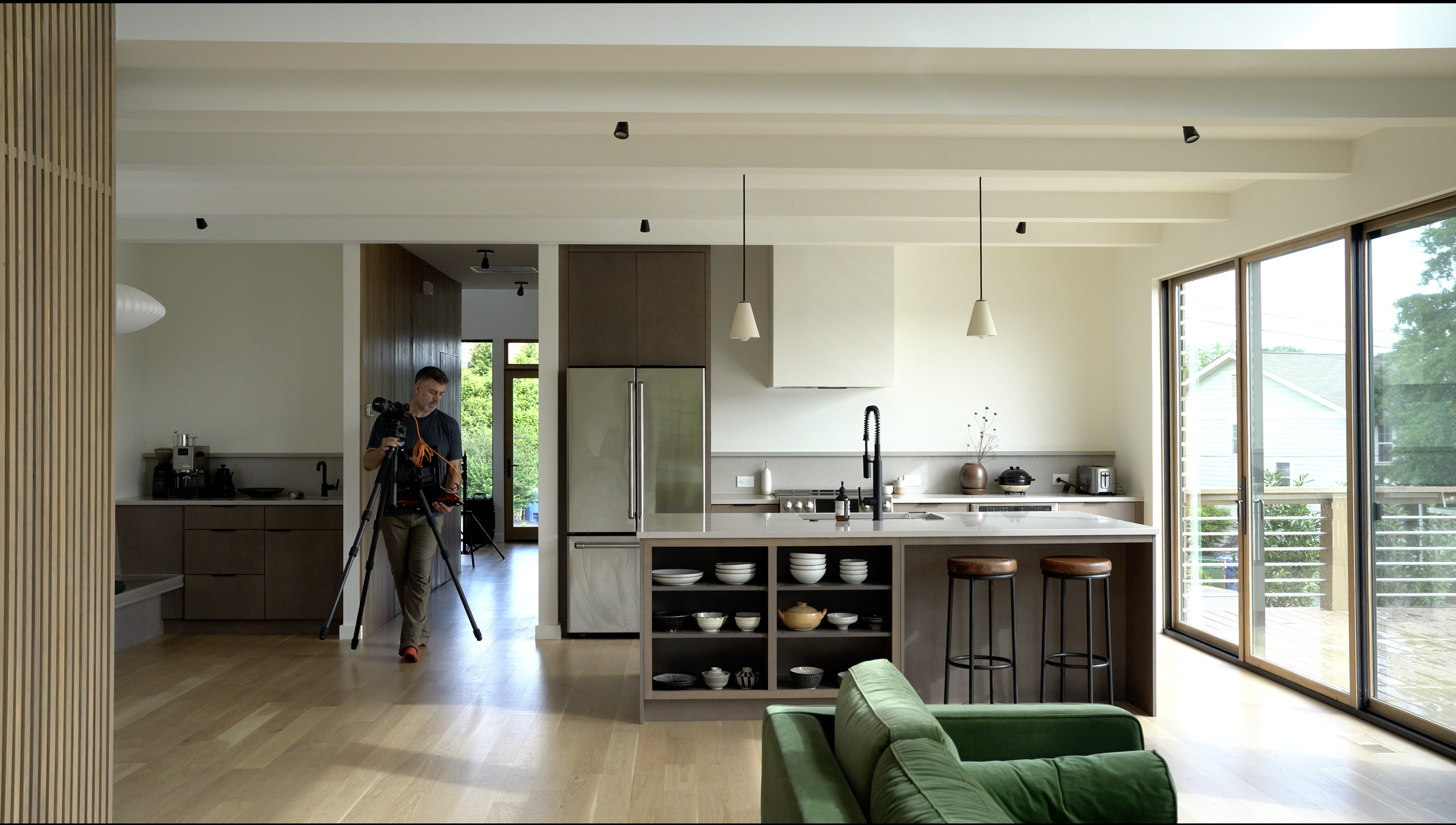 A man setting up camera equipment inside a modern kitchen with a large window and sliding glass door, natural light illuminating the space, with kitchen appliances and open shelving visible.