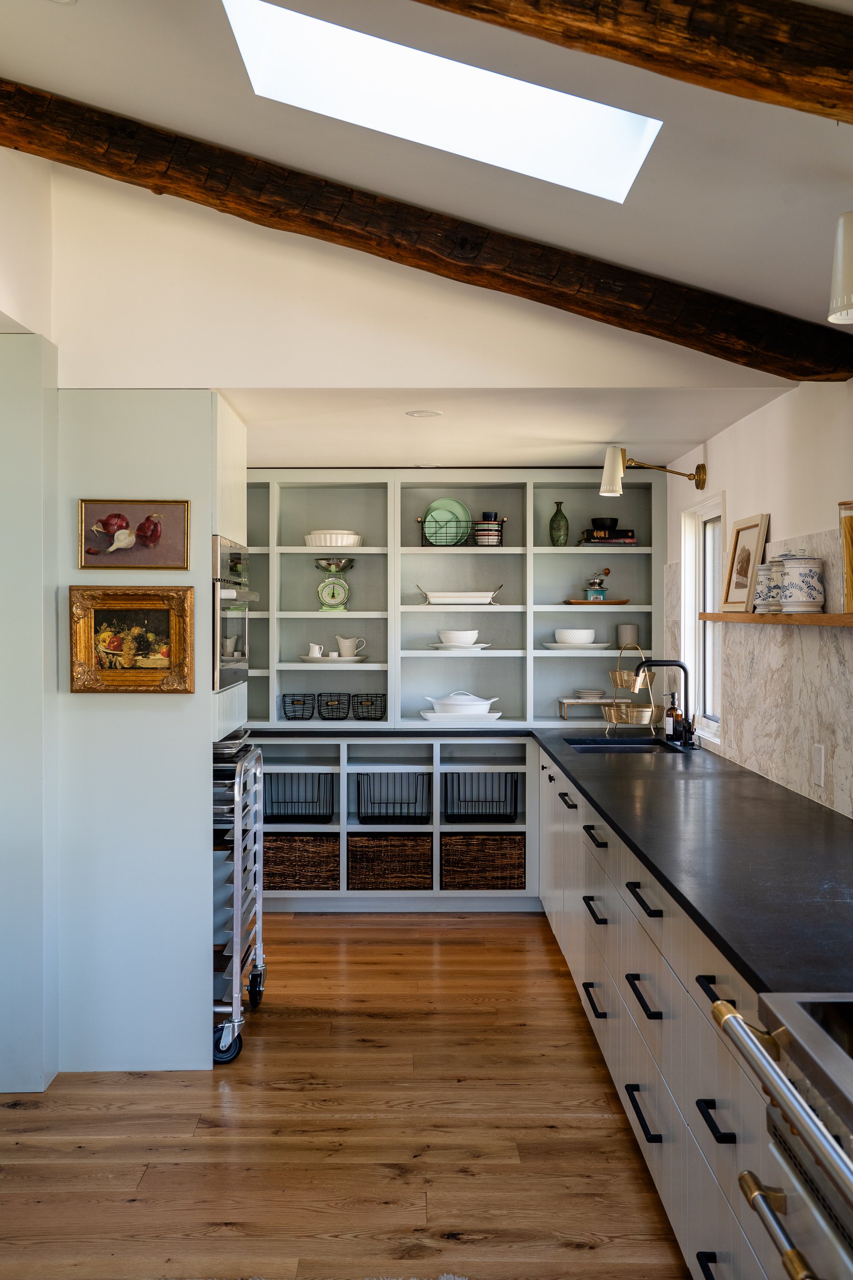 Kitchen pantry area with open shelving, black countertops, pale cabinetry, skylight, and wood floors.
