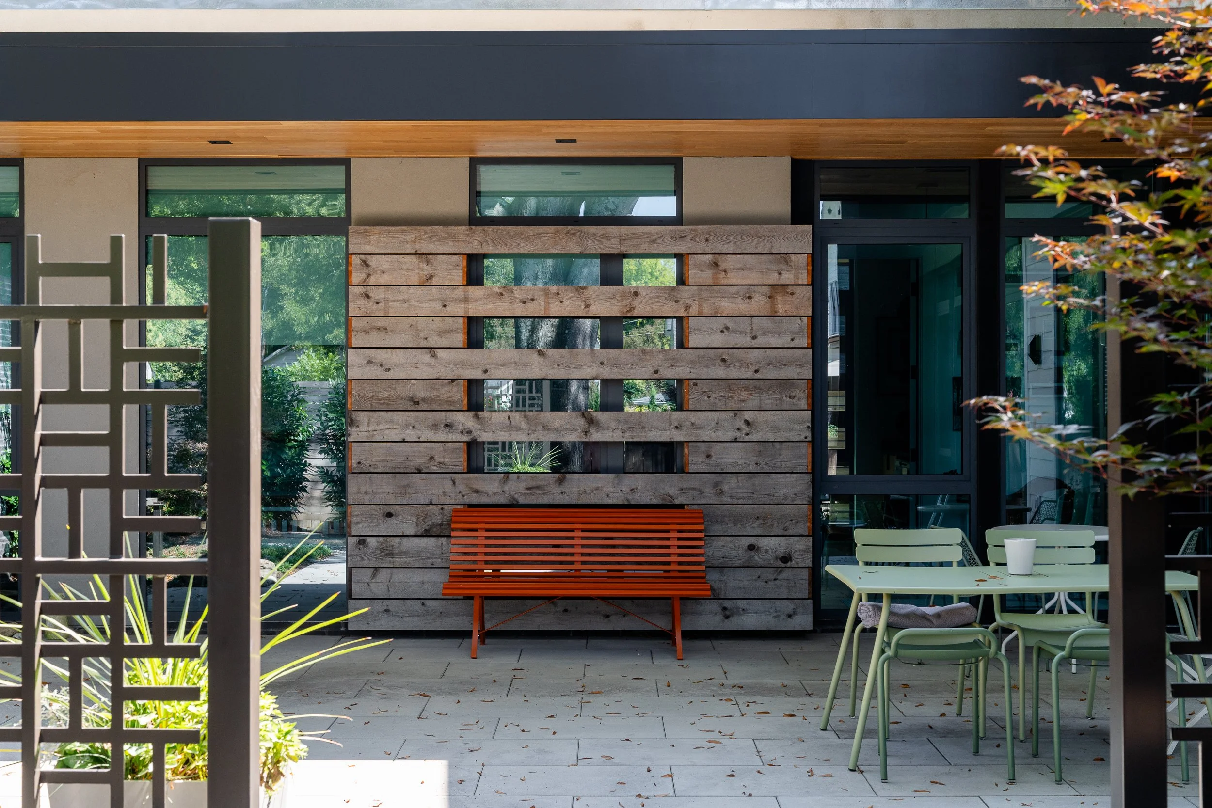An orange bench rests against a horizontal wood screen beside a glazed doorway and outdoor table beneath a roof overhang.