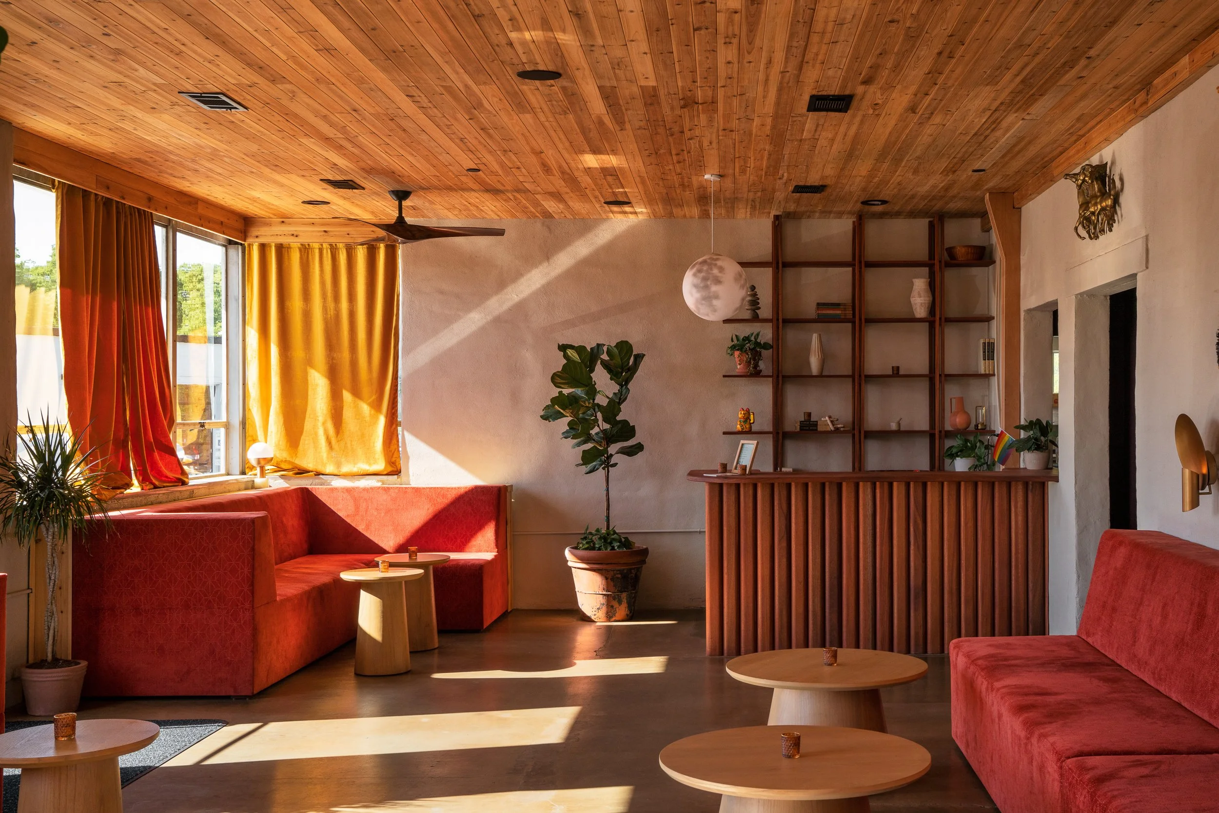 Interior of a living room with wooden ceiling and beams, red upholstered furniture, potted plants, yellow curtains, open windows, and decorative items on shelves.
