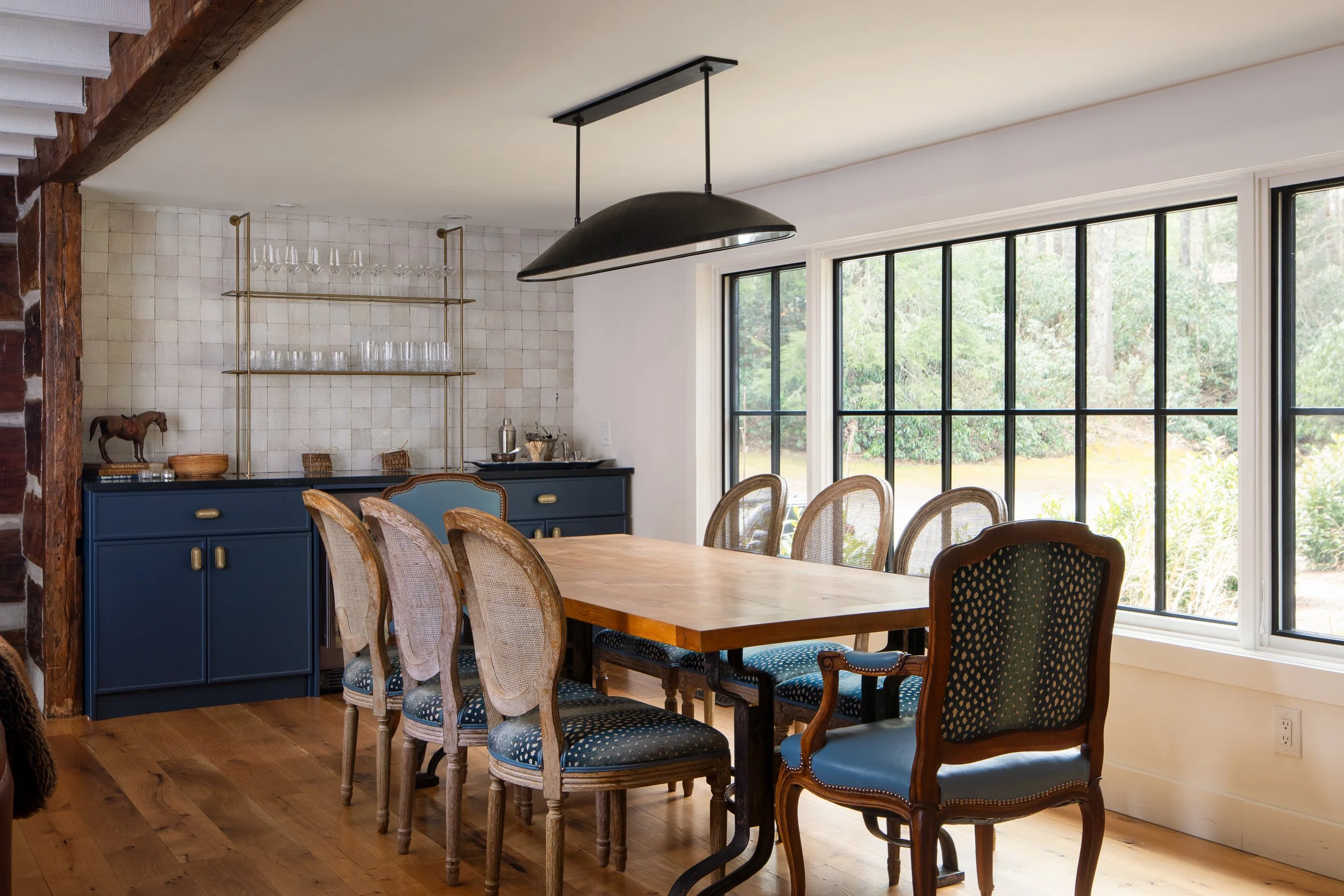 Dining room with mixed chairs, blue cabinet, tiled bar wall, large windows, and a black pendant light.