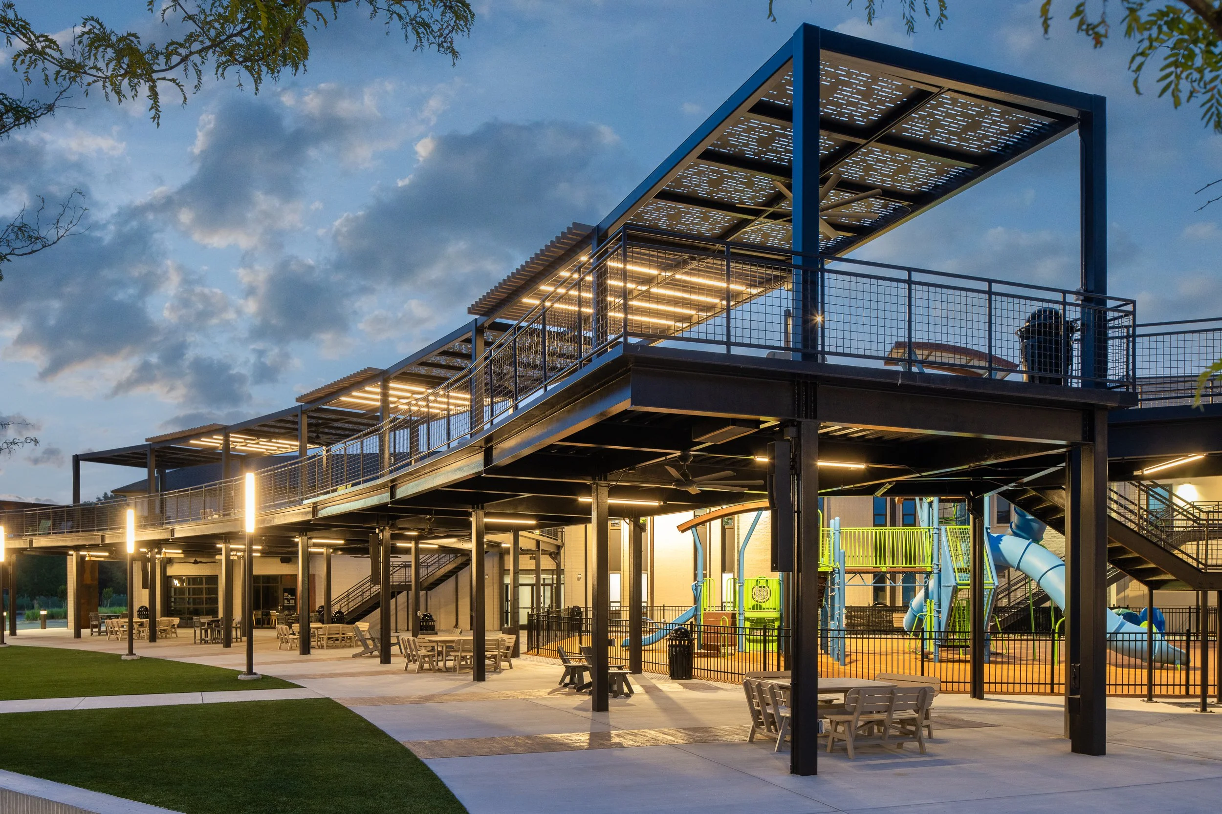 A modern playground structure with slides and climbing areas, illuminated by lights, under a blue sky with clouds at dusk, surrounded by a grassy field and seating area.