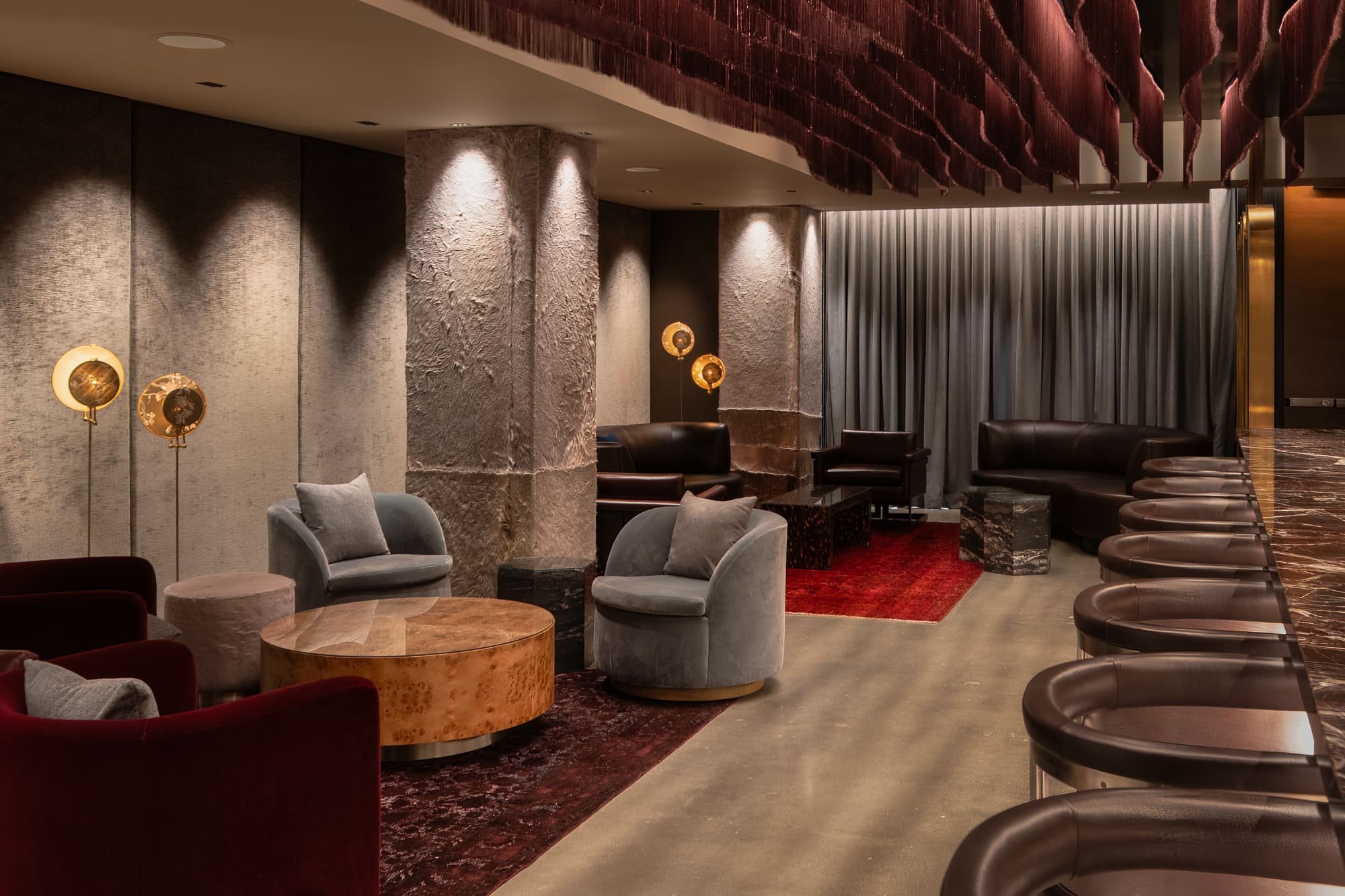 Hospitality lounge with velvet chairs, leather seating, textured columns, red rugs, and a sculptural burgundy ceiling installation.