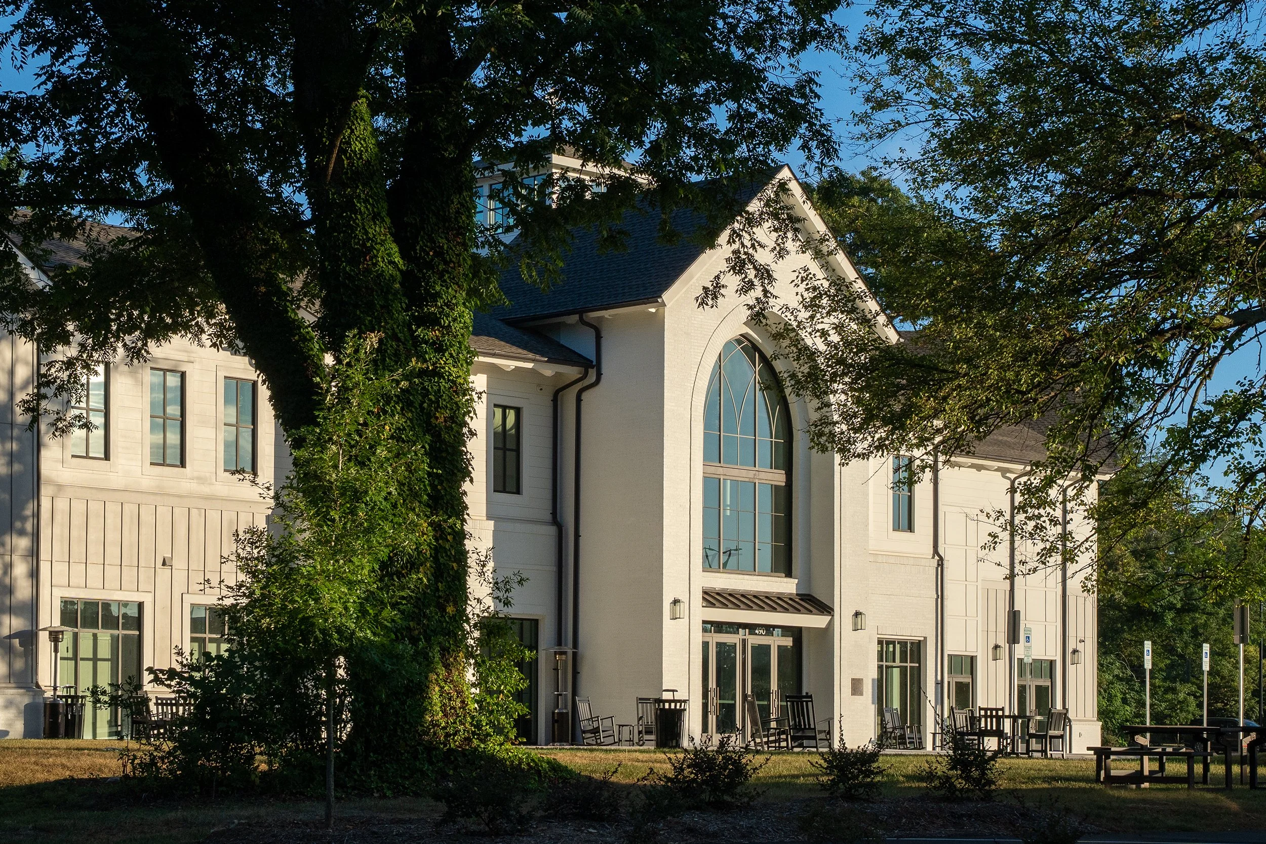 White brick exterior of Storyhill Church in Davidson, NC with a gothic arched window above the main entrance, framed by mature trees in morning light.