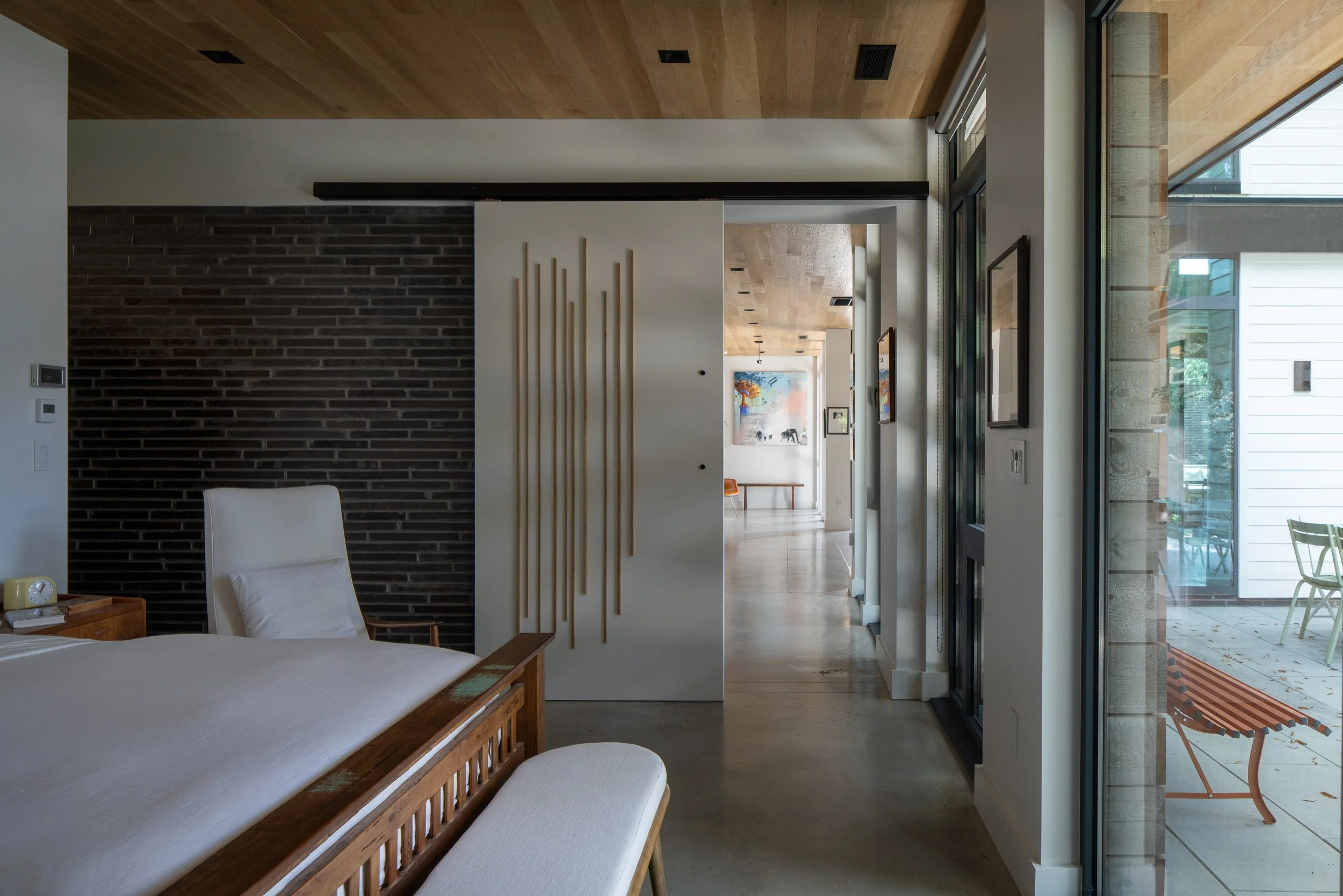 A bedroom with a dark brick wall connects to a bathroom through an open doorway beside a vertical wood screen and courtyard glazing.