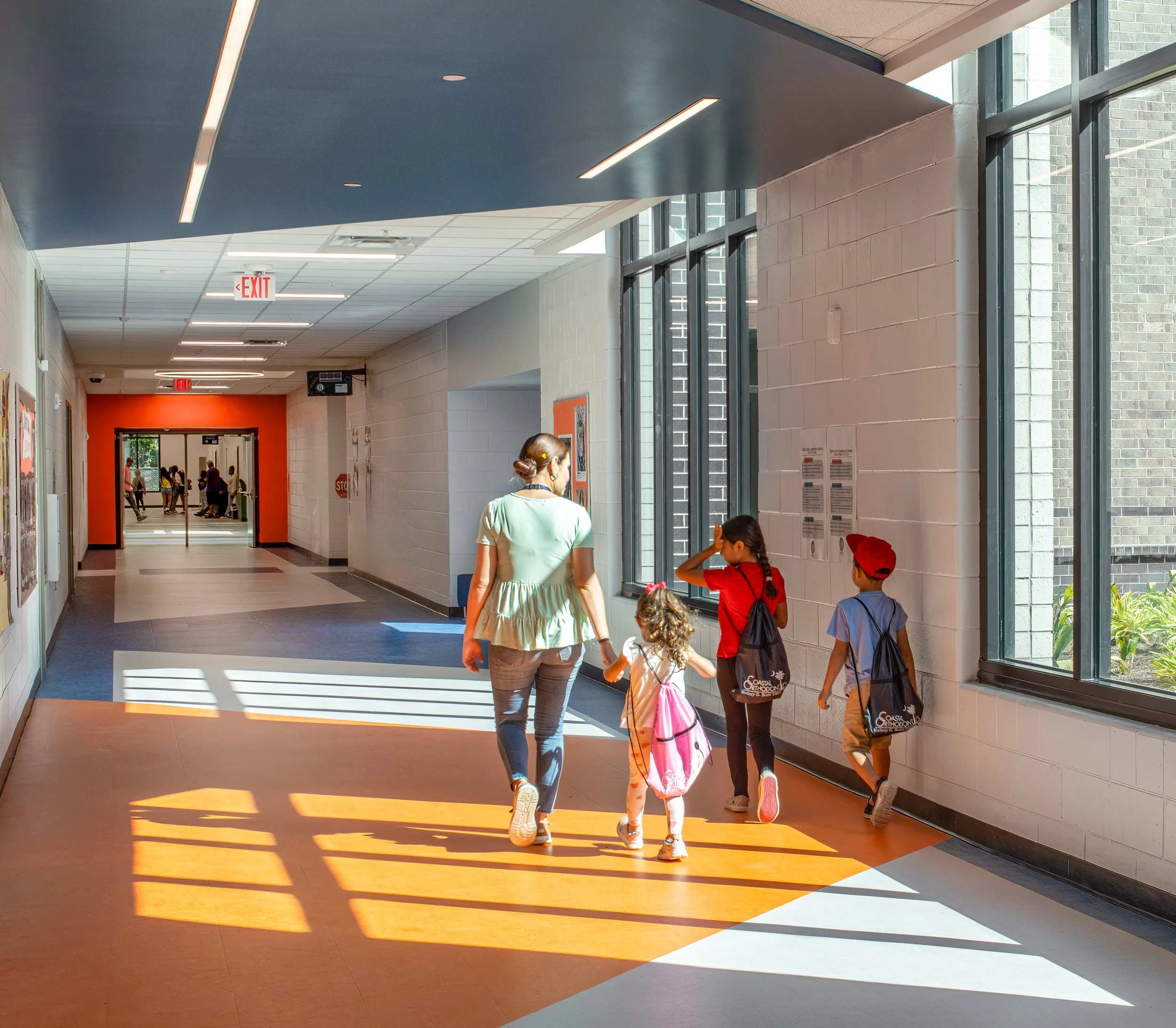 School corridor with students and an adult walking through bands of sunlight, with orange and blue design accents along the hallway.