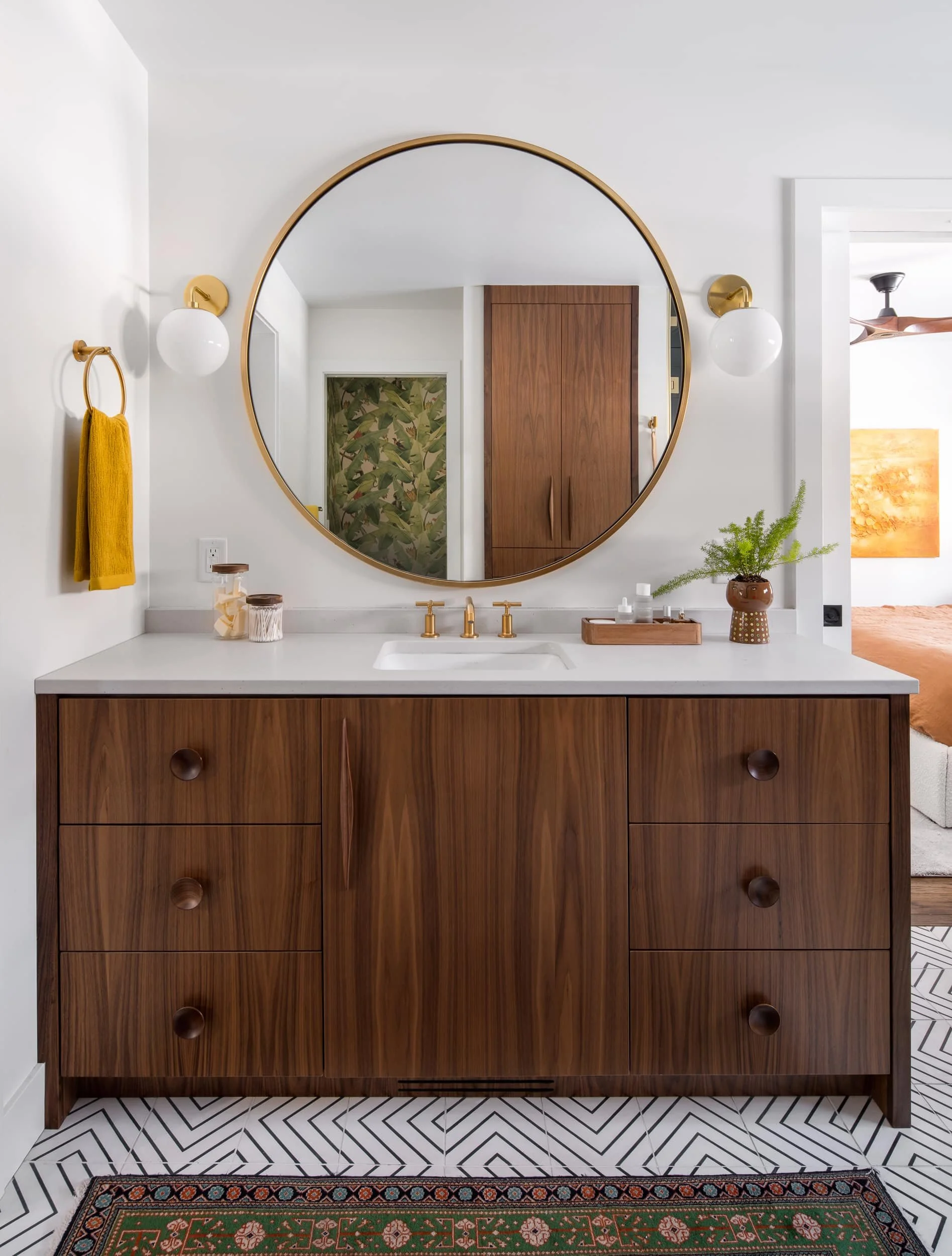 Primary bathroom vanity with walnut cabinetry, round brass-framed mirror, globe sconces, brass fixtures, geometric tile floor, and a view into the bedroom beyond