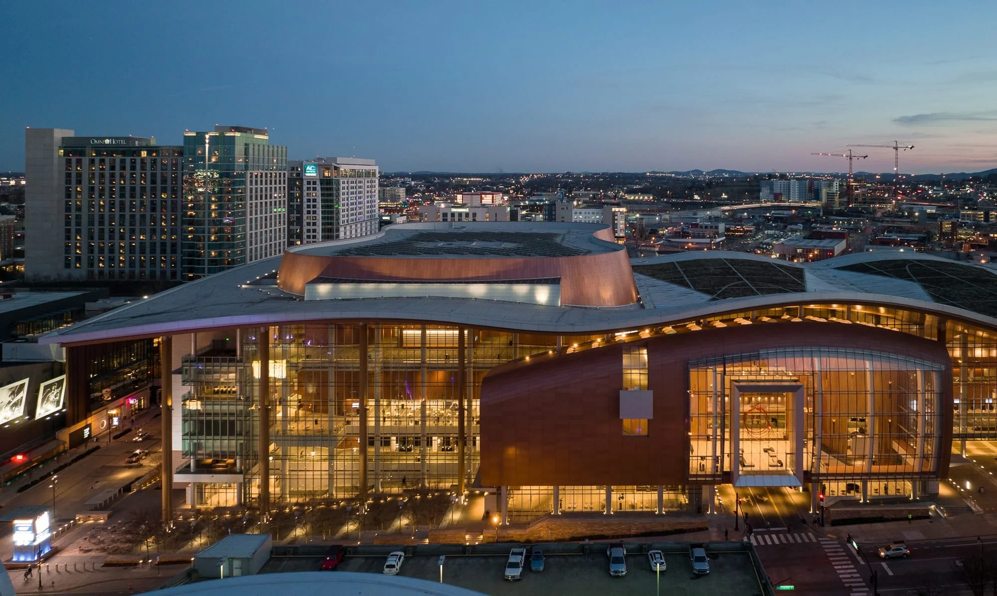 Aerial view of Music City Center in Nashville at dusk showing the sweeping curved copper facade, illuminated glass entry, green roof, and surrounding downtown hotels including the Omni and AC