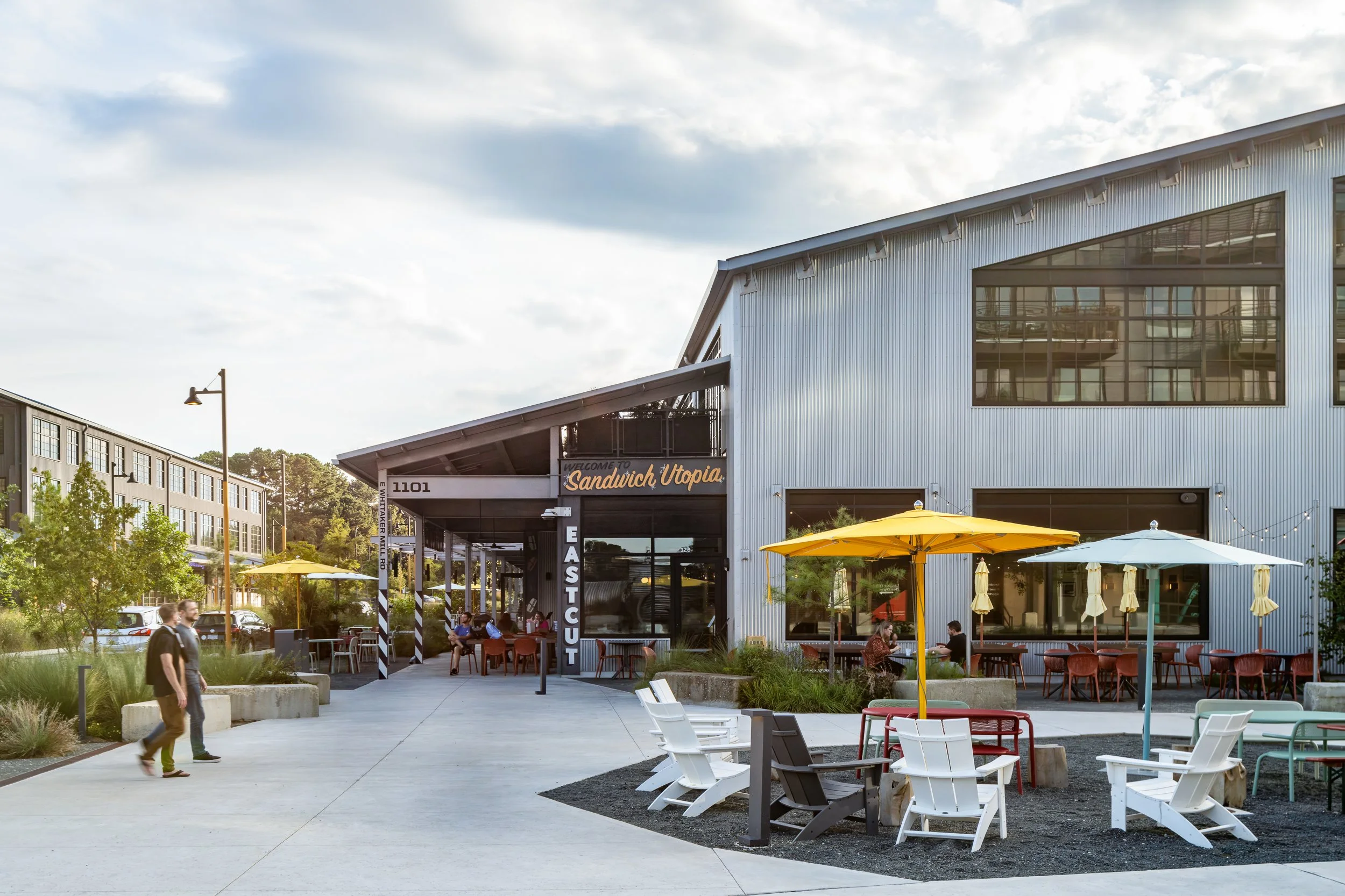 Plaza outside the Eastcut storefront with a Welcome to Sandwich Utopia sign, white Adirondack chairs around a fire pit area, yellow market umbrellas, and visitors walking through the space.