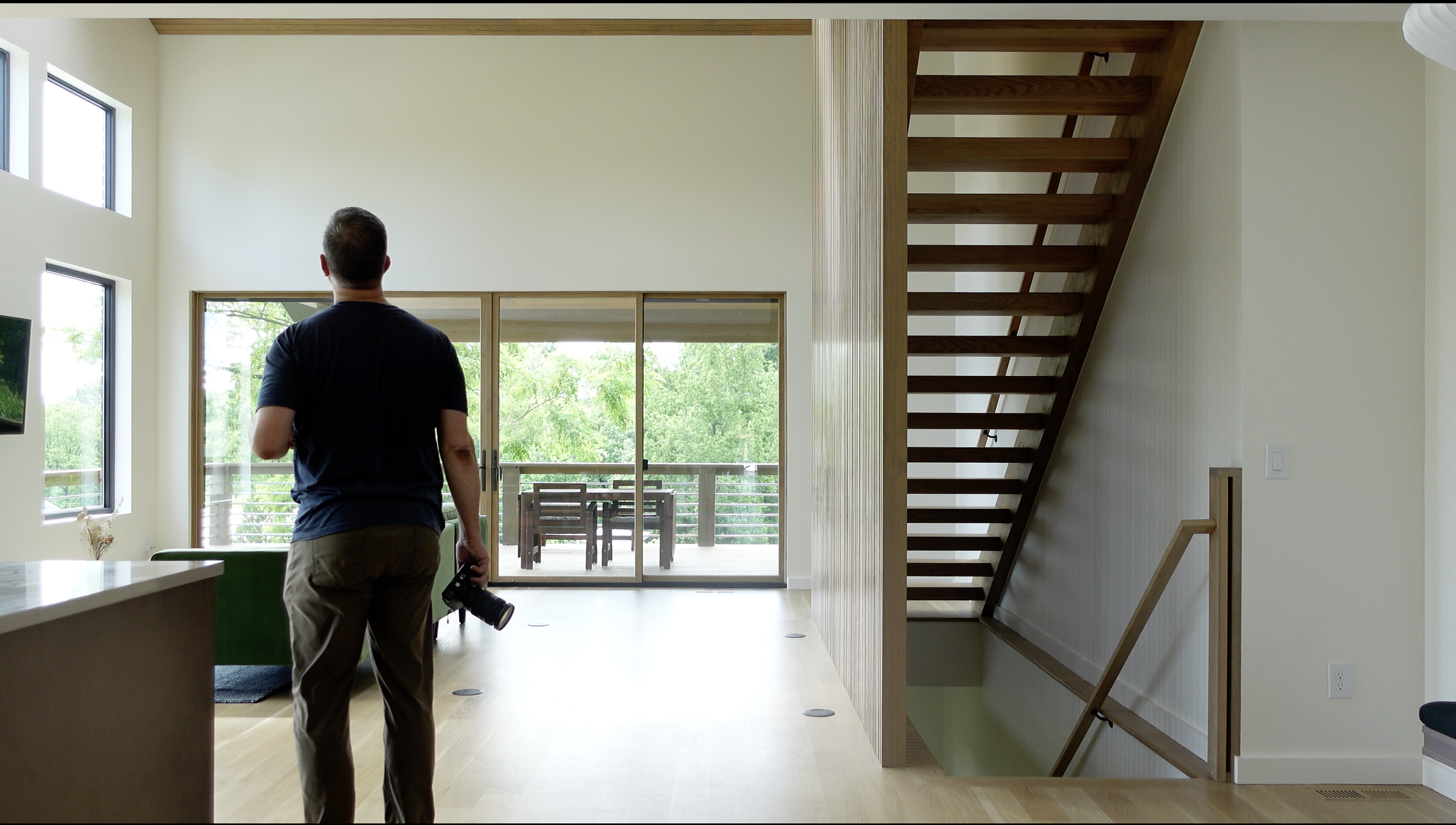 Man holding a camera walking inside a modern house, with sliding glass doors leading to a balcony, a staircase, and large windows letting in natural light.