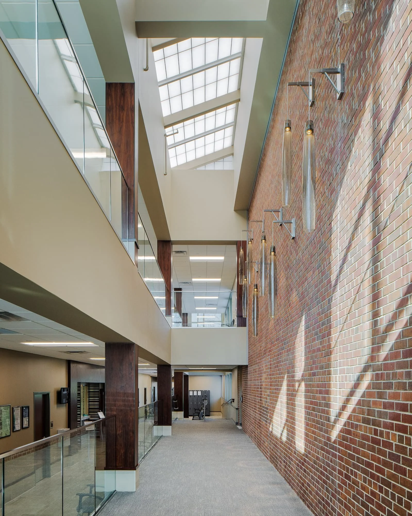 Multi-level interior corridor with brick feature wall, skylights, glass railing, and modern wall sconces.