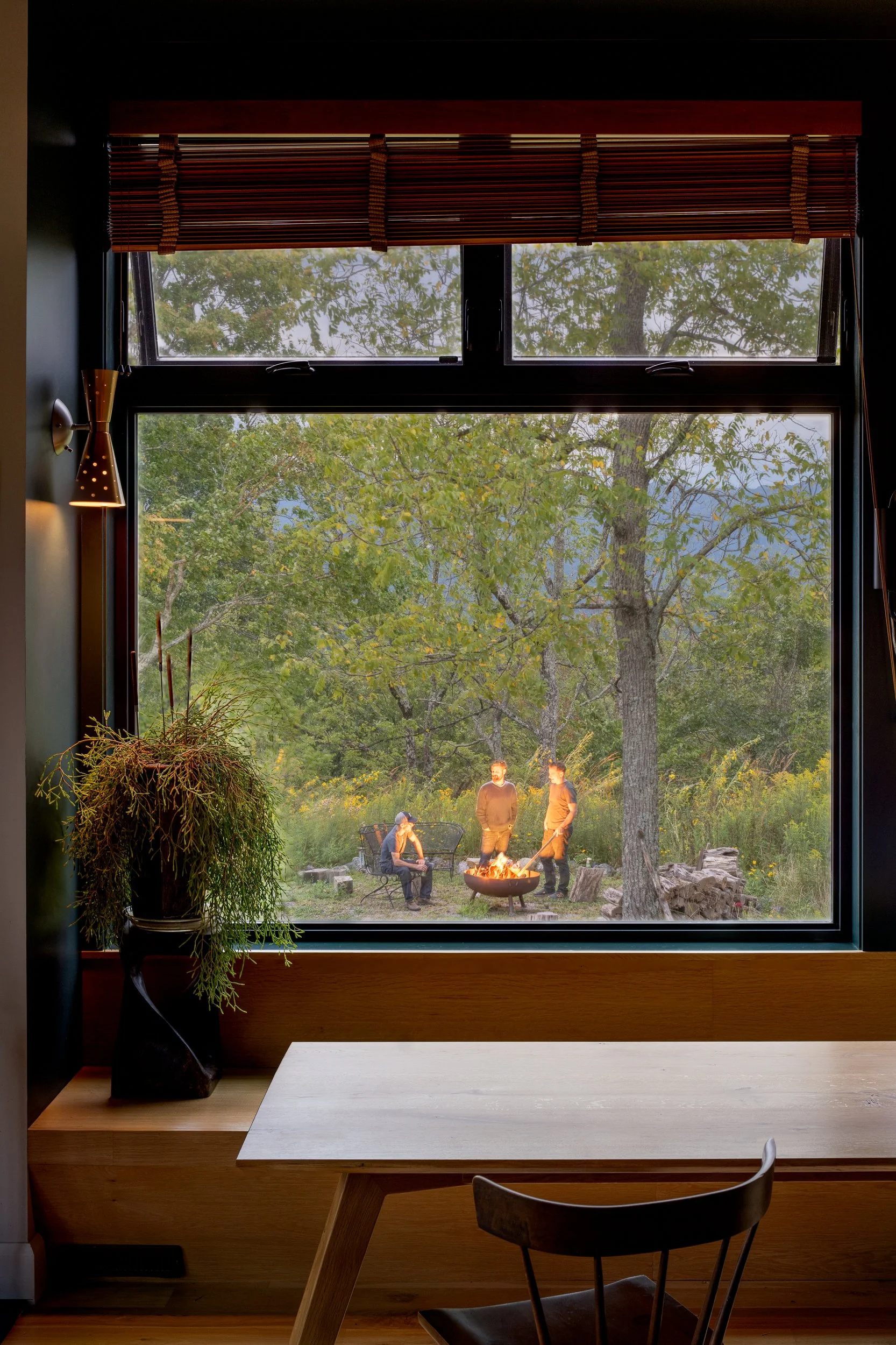 Large interior window framing people gathered around an outdoor fire pit at dusk.