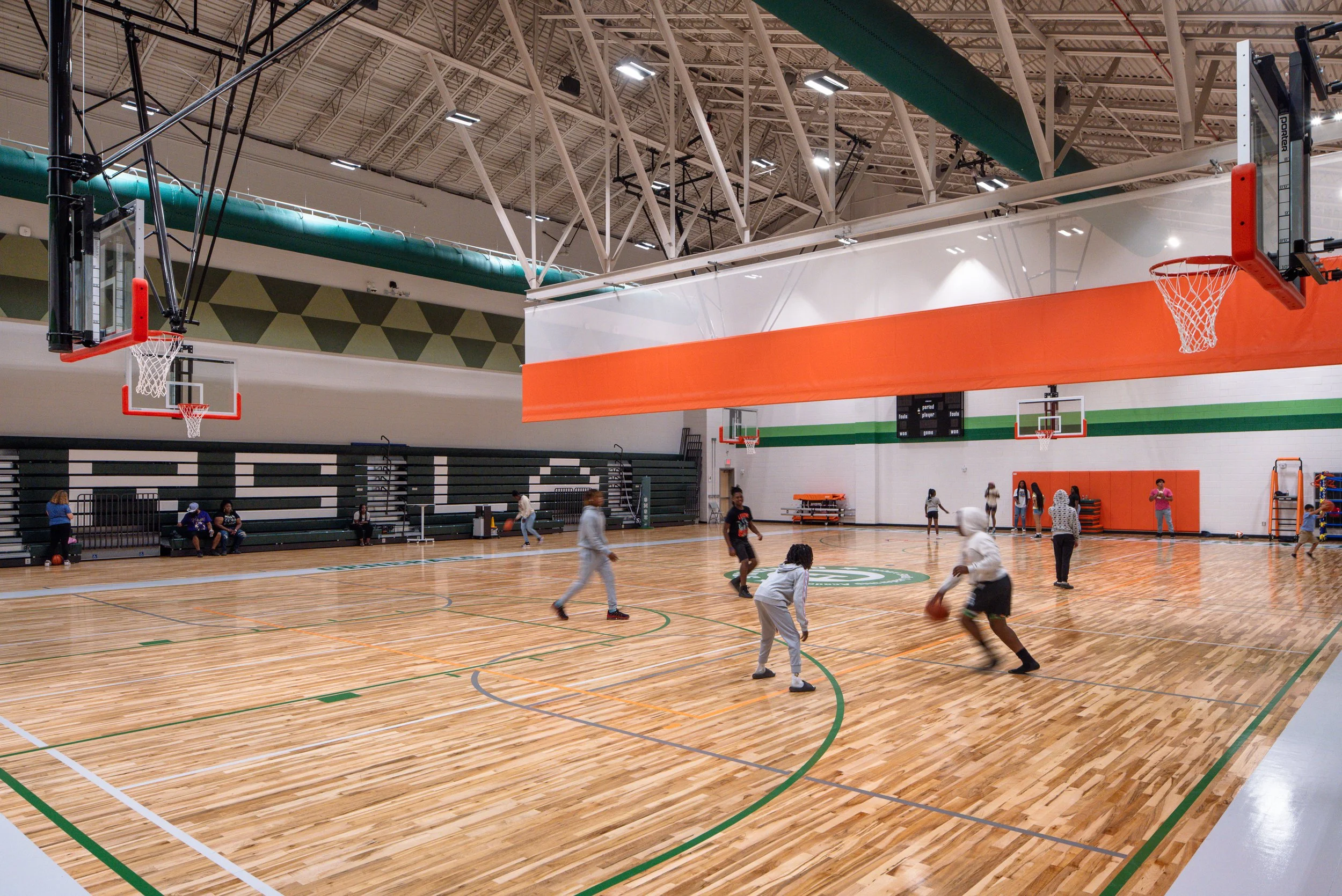 Large school gymnasium with polished wood floor, basketball hoops, exposed ceiling structure, and students playing on the court.