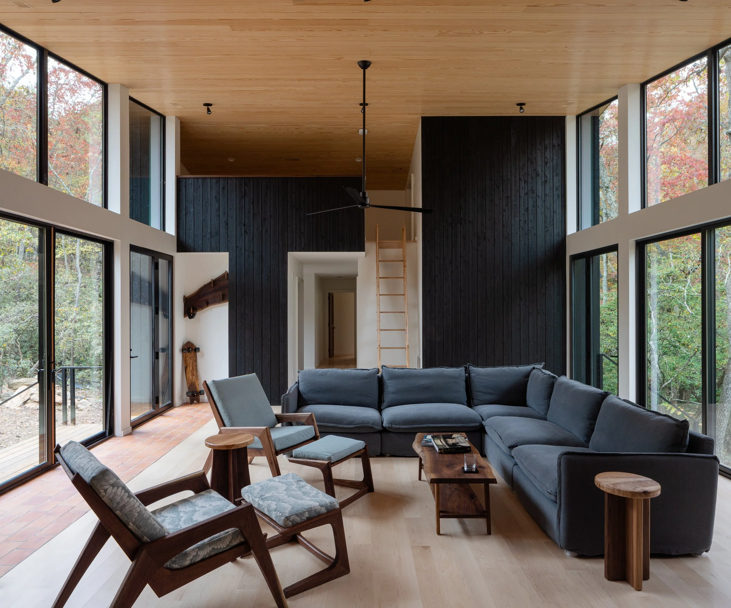 Living room with a tall wood ceiling, burnt cedar feature wall, large windows, and forest views on both sides.
