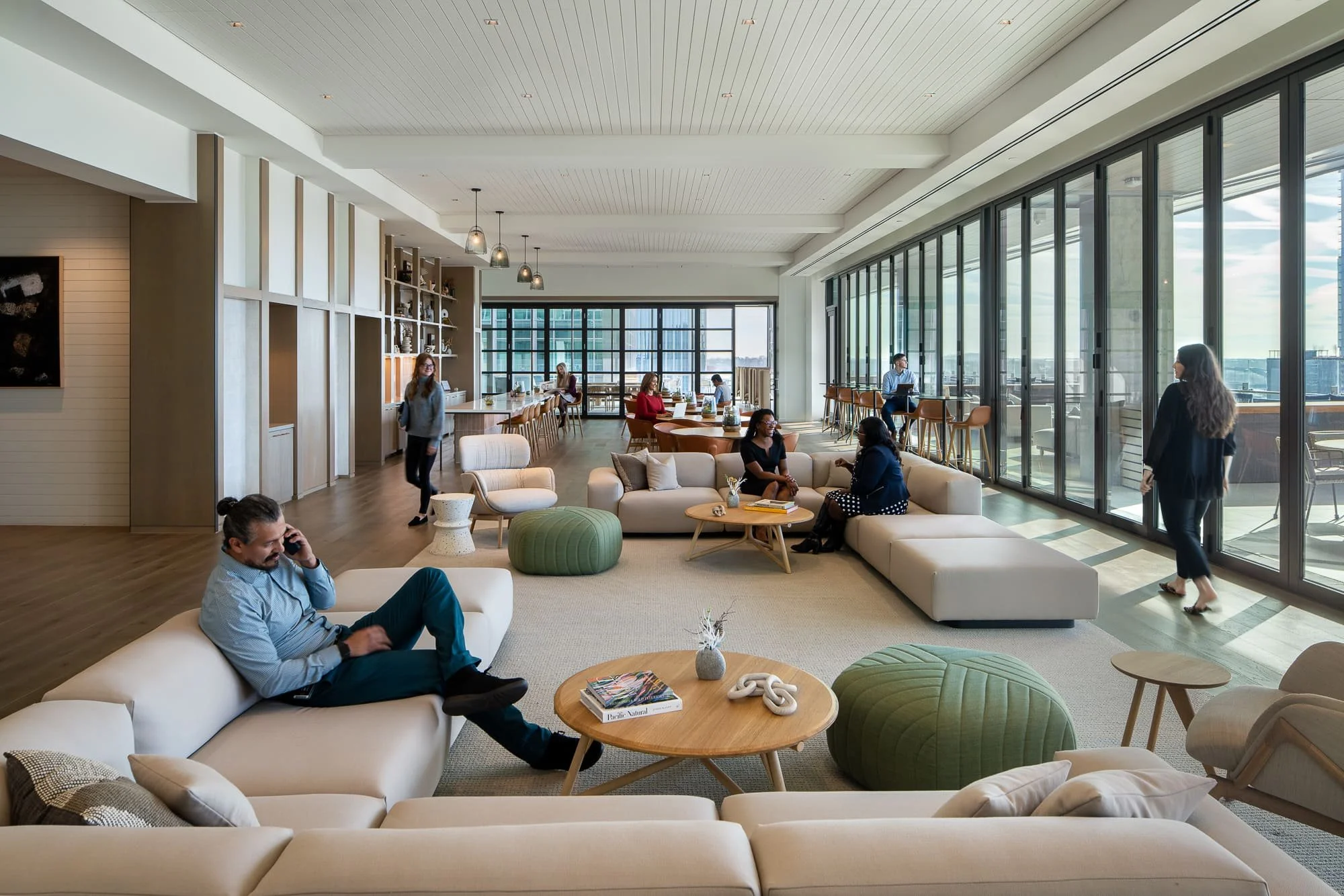 Modern amenity lounge with sofas, round tables, large windows, and people relaxing indoors.