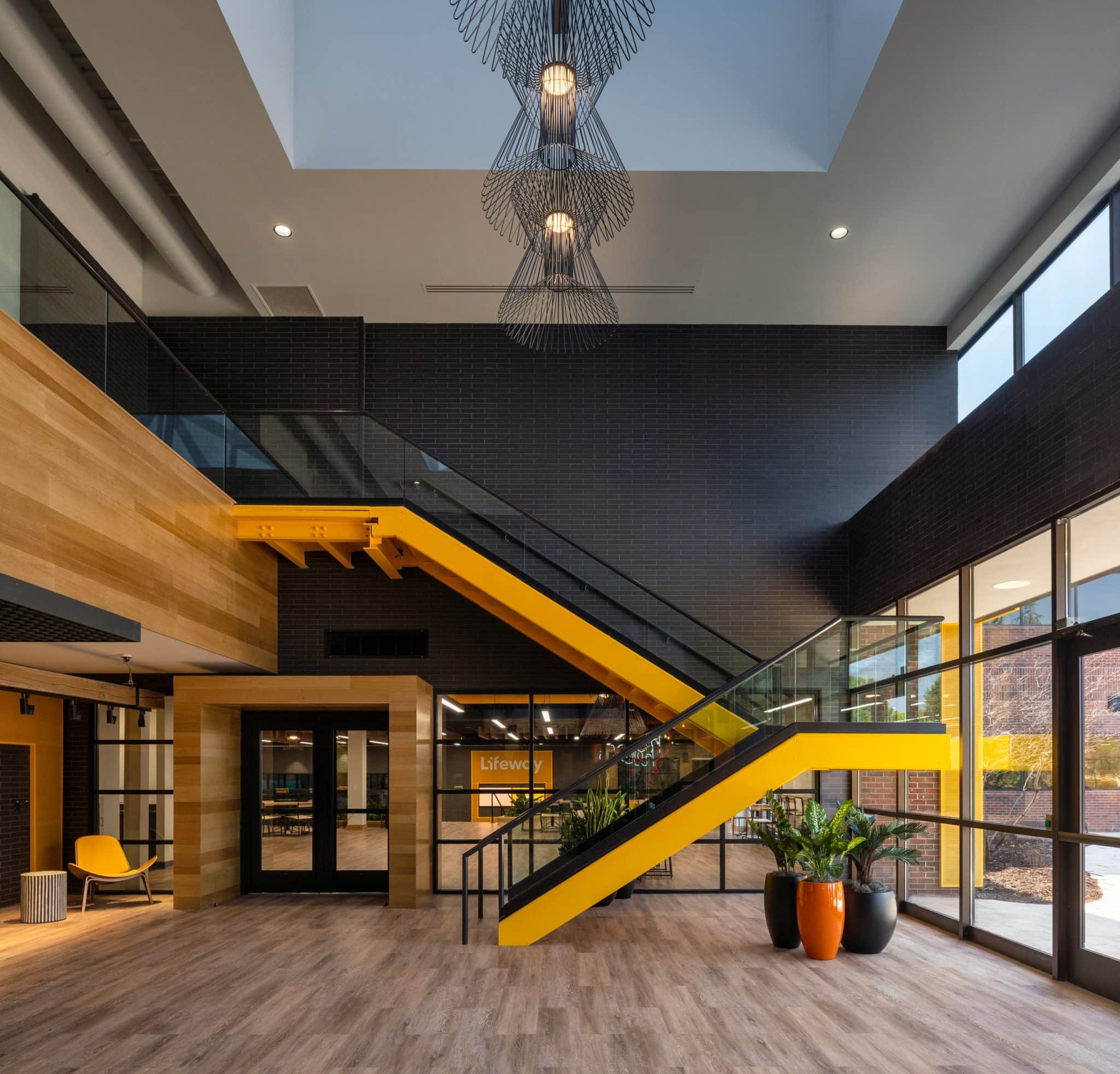 Modern office atrium with a bright yellow steel stair, black brick walls, wood cladding, glass railings, and tall windows.